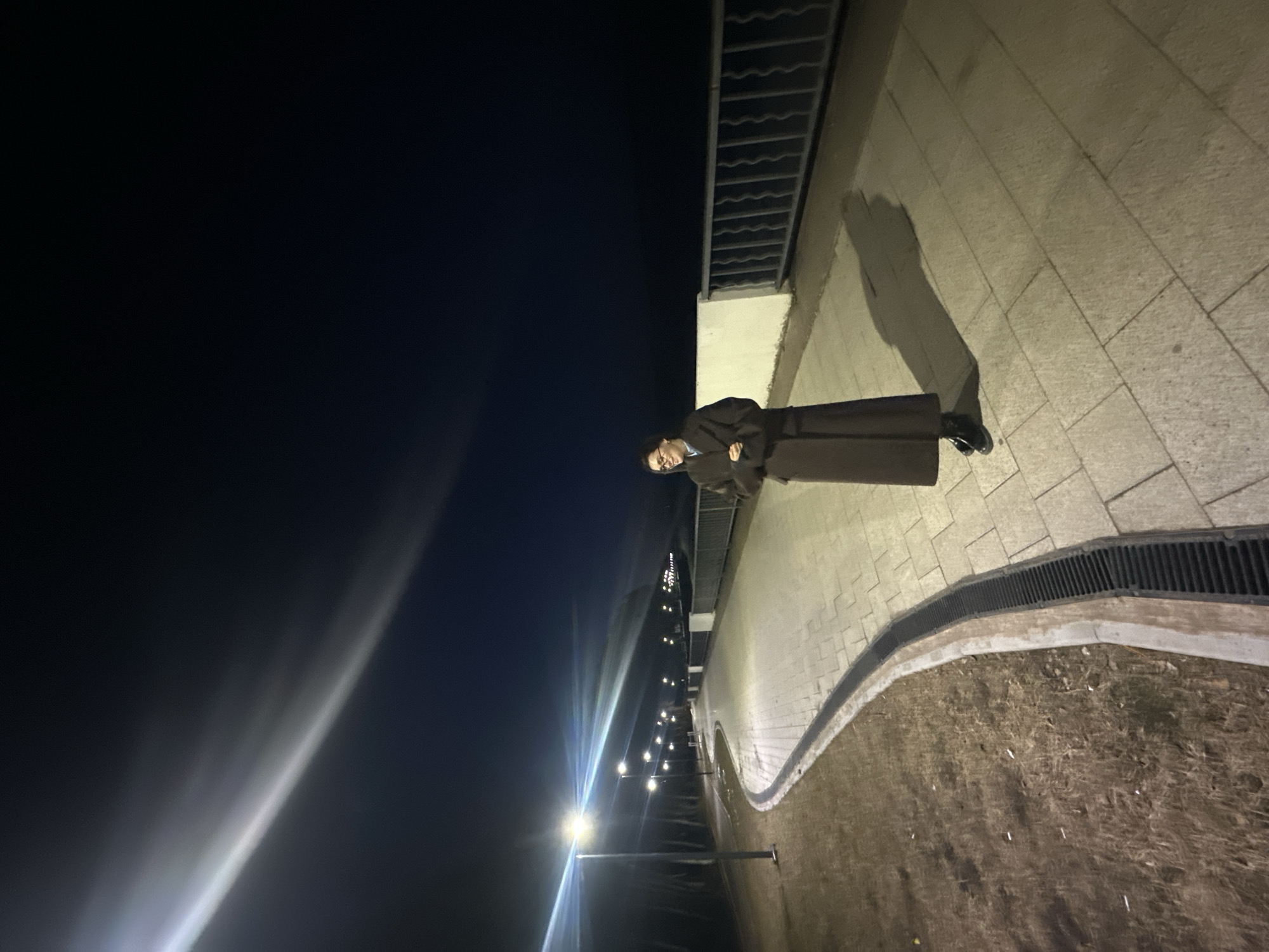 A person stands on a walkway in Chaikovsky, Russia at night. The person wears a dark, long coat and dark shoes, casting a long shadow. The walkway is made of light-colored paving stones and bordered by a metal railing and a grassy area. Streetlights illuminate the walkway and the night sky is dark.