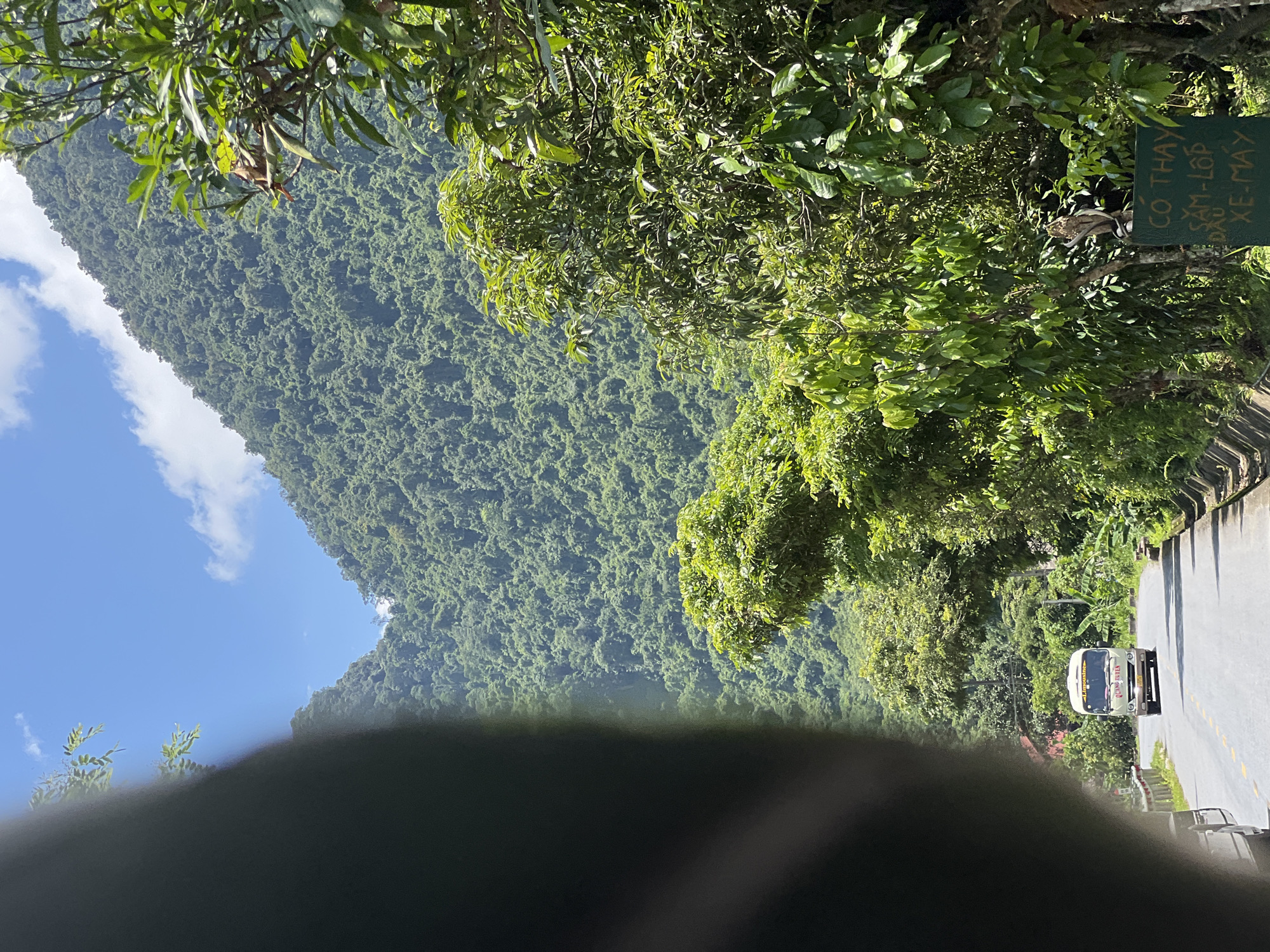 The image shows a road winding through a lush, mountainous landscape in Thuận Hòa Commune, Vietnam. The road is paved and has yellow lines marking the lanes. A white bus with "LIMOUSINE" written on it is driving away from the camera. The mountainsides are covered in dense green vegetation. Trees line the side of the road, and a green sign with Vietnamese text is visible on the right. The sky is blue with a few white clouds. A dark, blurry object, likely a part of the vehicle the photo was taken from, occupies the left foreground.