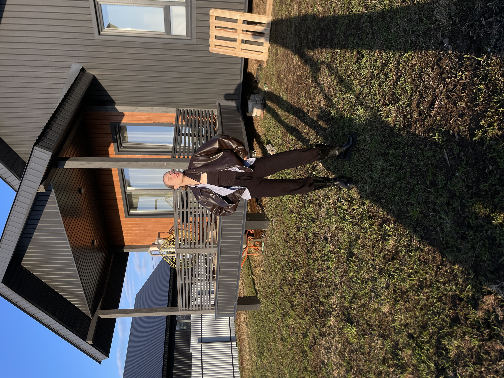 A young person, appearing to be female, stands outdoors in Podshivalovo, Russia. She is the central subject, dressed in a dark brown leather jacket over a light-colored collared shirt and dark trousers, complemented by black boots. Her hair is pulled back, and she is captured in a posed stance, leaning slightly backward with her head tilted upwards, seemingly looking towards the sun.
The setting features a modern-style house with dark grey horizontal siding and contrasting wooden paneling around windows and on a porch structure. Several windows reflect the clear blue sky. The ground is a mix of sparse green grass and patches of bare earth. To the right of the person, a wooden pallet rests on the ground. Distinct, long shadows cast by the person and the house indicate bright, low sunlight. The house has a covered porch or overhang supported by dark beams and a railing. The sky is visibly clear and blue in the upper left of the image.
The activity taking place is primarily posing for a photograph, with the subject seemingly enjoying the sun's warmth. The time of day is suggested to be either early morning or late afternoon due to the strong, elongated shadows. The weather appears fair and sunny. There is no visible text in the image.