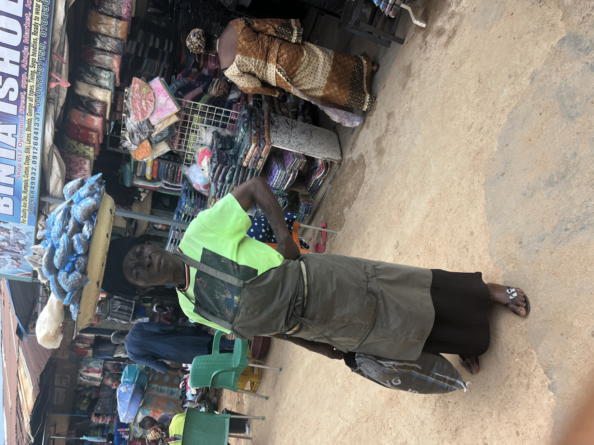 A medium shot shows a woman standing in a market stall in Jos, Nigeria. The woman is wearing a lime green shirt and a grey apron over a black skirt, and she is carrying a dark-colored bag. She is looking toward the camera with a serious expression. In the background, various market goods, including textiles and bags of food, are displayed. Two other women and a man are visible in the background, also engaged in market activities. The ground is made of dirt and concrete, and the stalls are covered with corrugated metal roofs. The overall scene depicts a bustling marketplace environment.