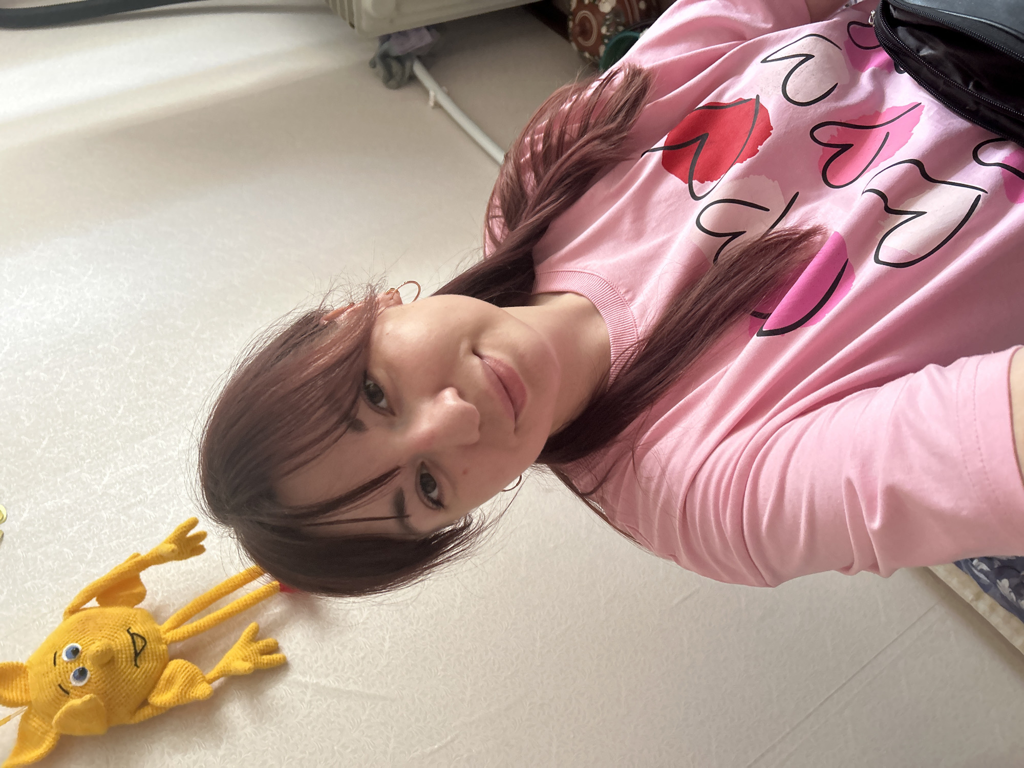 A young woman with reddish-brown hair styled in pigtails and bangs is taking a selfie indoors. She is wearing a pink t-shirt adorned with heart designs in shades of red and pink. To her upper left, a yellow crocheted doll is attached to a wall with a textured pattern. The background shows a plain, light-colored wall and a radiator. There is a black bag visible at the lower right of the frame. The scene appears to be a domestic setting within Krasnovishersk, Russia.