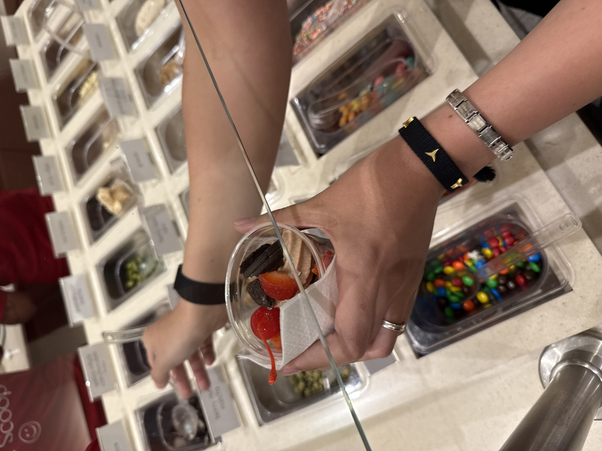 A close-up, high-angle indoor shot depicts a person preparing a dessert at a self-serve frozen yogurt shop. The person is adding toppings to a clear plastic cup filled with frozen yogurt, including Oreo cookies, strawberries, and a maraschino cherry. The cup is held by one hand with a ring on its finger, while the other hand is reaching towards various topping containers. The person is wearing two bracelets on their left wrist: a black woven bracelet with a gold emblem and a silver, segmented bracelet. In the background, a variety of toppings are visible in clear plastic containers, including sprinkles, M\&Ms, and other dessert items. The containers are arranged in a grid-like display, with labels indicating the contents of each. The environment appears clean and well-lit, typical of a frozen yogurt shop. Based on the "Scoop" logo in the bottom left corner and the overall layout, the location is likely in Mexico City, Mexico.