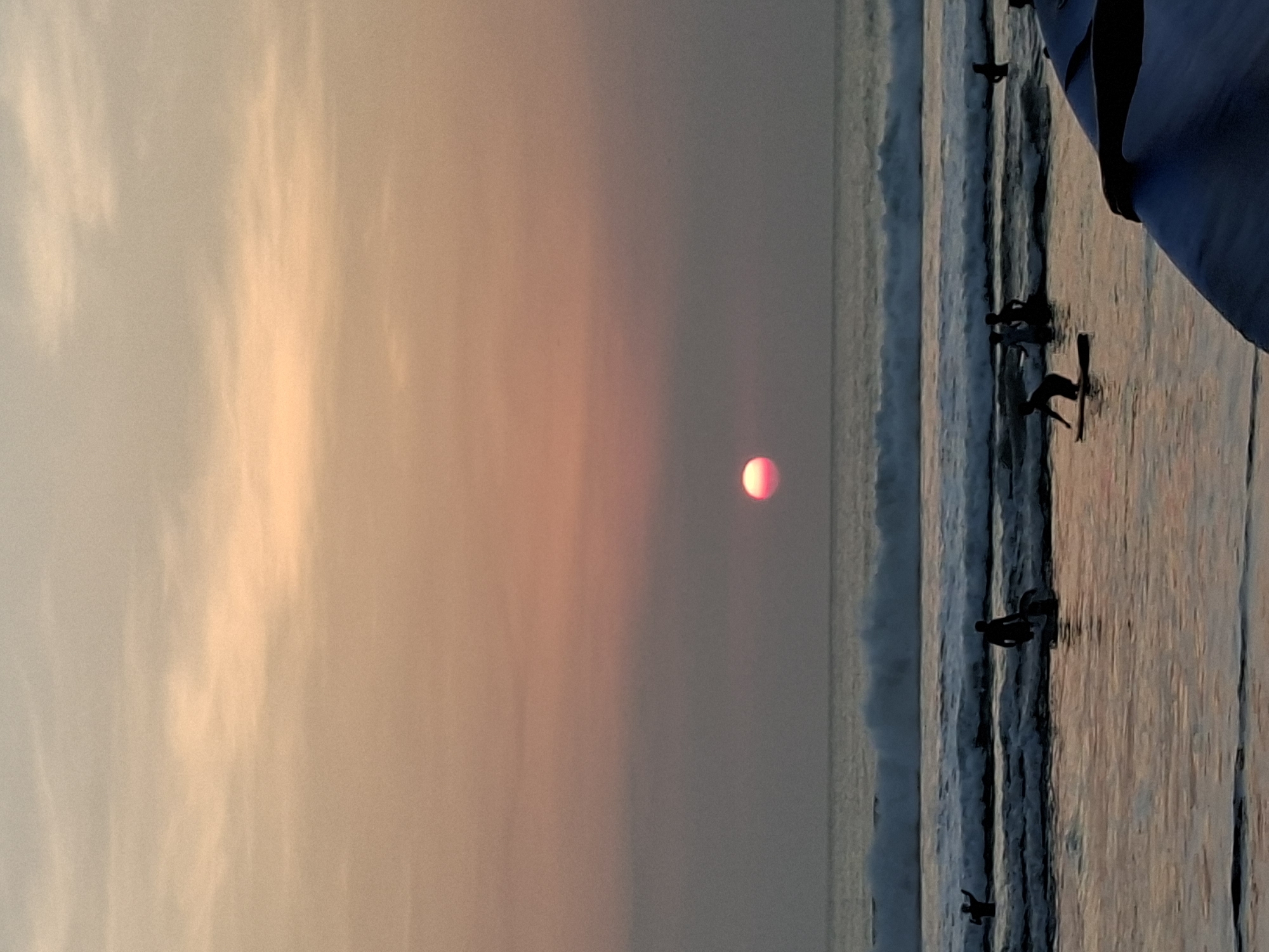 This photograph captures a sunset scene at Kuta Beach, Bali, Indonesia. The sun, a vivid pink orb, is partially obscured by the horizon, casting a warm glow across the sky and reflecting on the water. Gentle waves roll towards the shore, where several surfers are visible. Some are standing on their surfboards, while others are in the water, preparing to catch a wave. The silhouettes of the surfers are stark against the colorful sky and the turbulent sea. The foreground is dominated by the edge of a surfboard, suggesting the photographer is also participating in the activity. The overall atmosphere is tranquil and serene, a typical evening at this popular surfing destination.