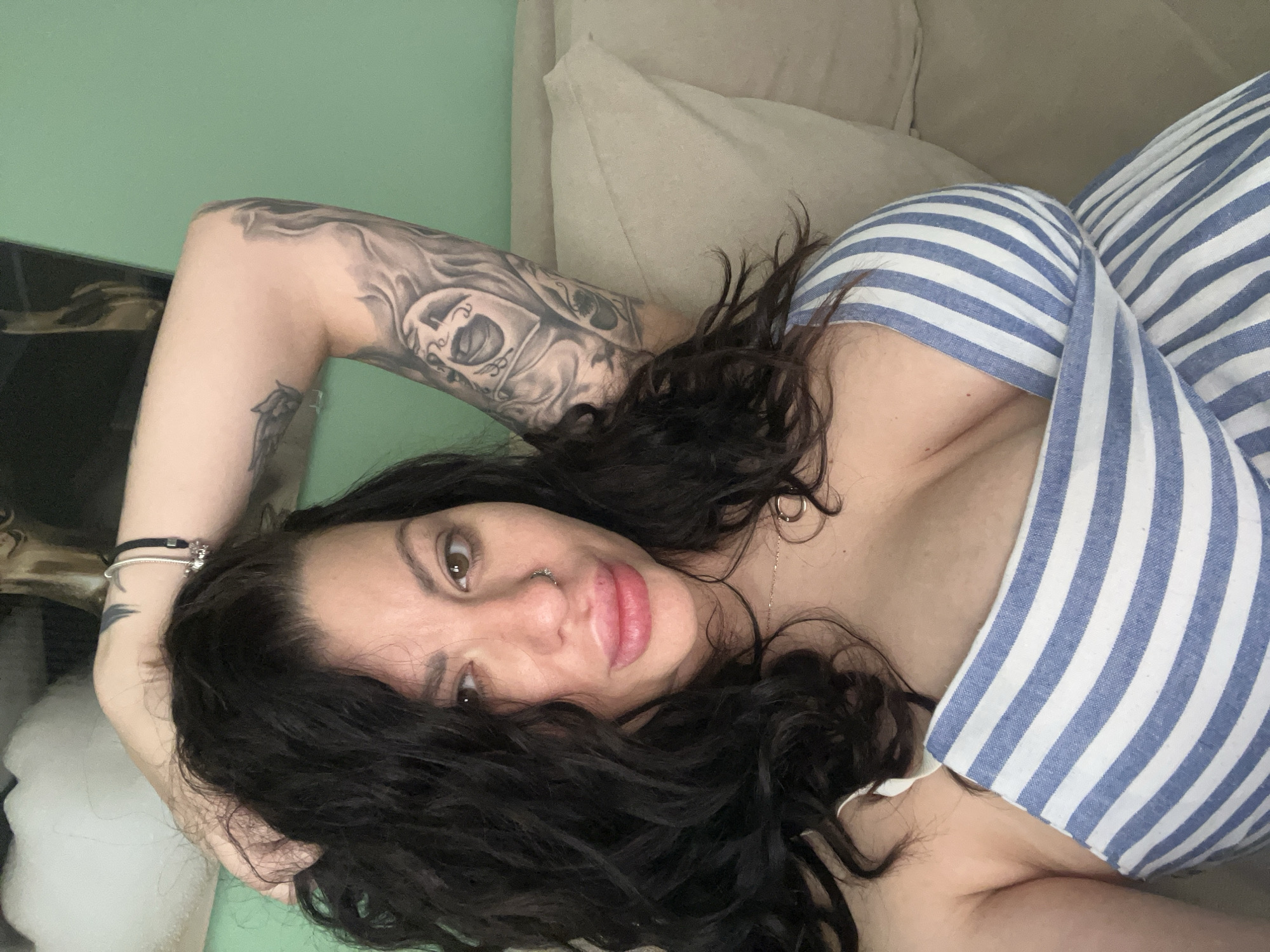 A woman with dark hair and tattoos on her left arm is reclining on a beige sofa against a light green wall. She is wearing a blue and white striped top. She has dark eyebrows, brown eyes, and a nose piercing. The location in Plovdiv, Bulgaria cannot be determined from the image.