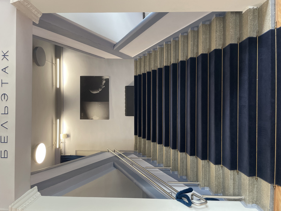 The image displays the interior of a grand staircase within the Bel Etage Concert Hall in Moscow, Russia. The staircase, constructed of light grey granite or stone, ascends directly towards the viewer in a symmetrical fashion. A dark blue carpet runner with thin golden horizontal trim lines covers the central portion of the steps. Stainless steel handrails with vertical support posts are present on the left side, with a blue velvet rope loosely looped around a lower section of the handrail. The walls flanking the staircase are white, accented by light grey architectural beams that define the stairwell.
At the top of the visible staircase, a landing features a low, dark-colored, rectangular bench or console table positioned centrally. Above this, a large horizontal black-and-white artwork is mounted on the wall. The artwork depicts two abstract forms; the right side clearly shows the profile of a bald head looking downwards in deep shadow, while the left side is more abstract.
The ceiling, painted white or light grey, is equipped with various lighting fixtures: a large circular flush-mount light in the upper left, and multiple linear fluorescent lights extending horizontally across the ceiling further back. The Russian word "БЕЛЬЭТАЖ" (Bel Etage), translated as "Dress Circle" or "First Floor," is prominently displayed in large, dark blue sans-serif letters across the upper ceiling area, indicating the venue's name or a specific section. No individuals are present in the scene. The overall environment appears well-maintained and designed for public events or performances.