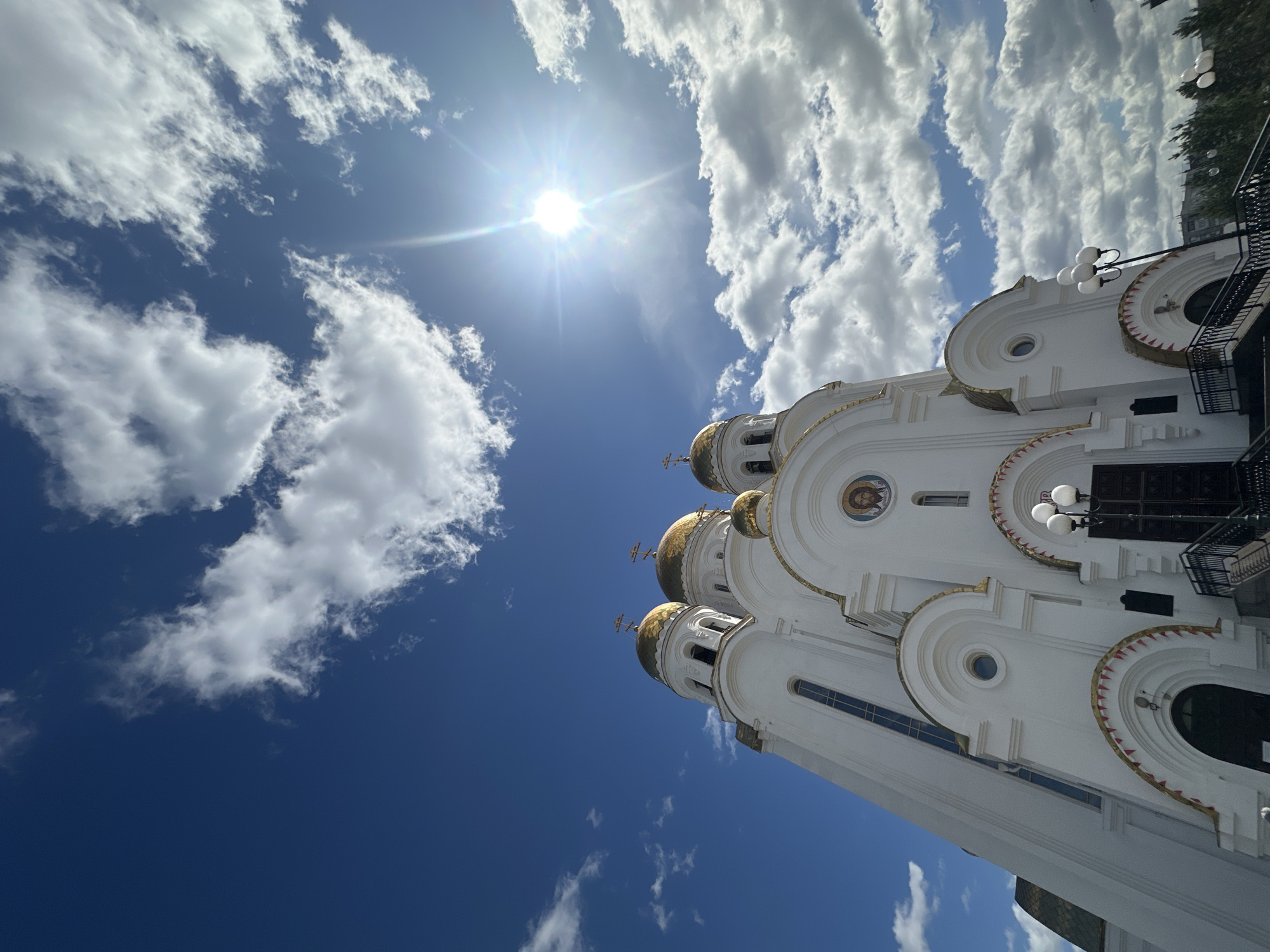 The image captures a low-angle view of the Cathedral on the Blood in Yekaterinburg, Russia, against a backdrop of a bright blue sky with scattered white clouds and the sun shining intensely. The cathedral's white facade with golden domes is prominently displayed, showcasing its ornate architectural details, including religious iconography and windows. The surrounding environment includes streetlights and glimpses of other buildings and greenery.