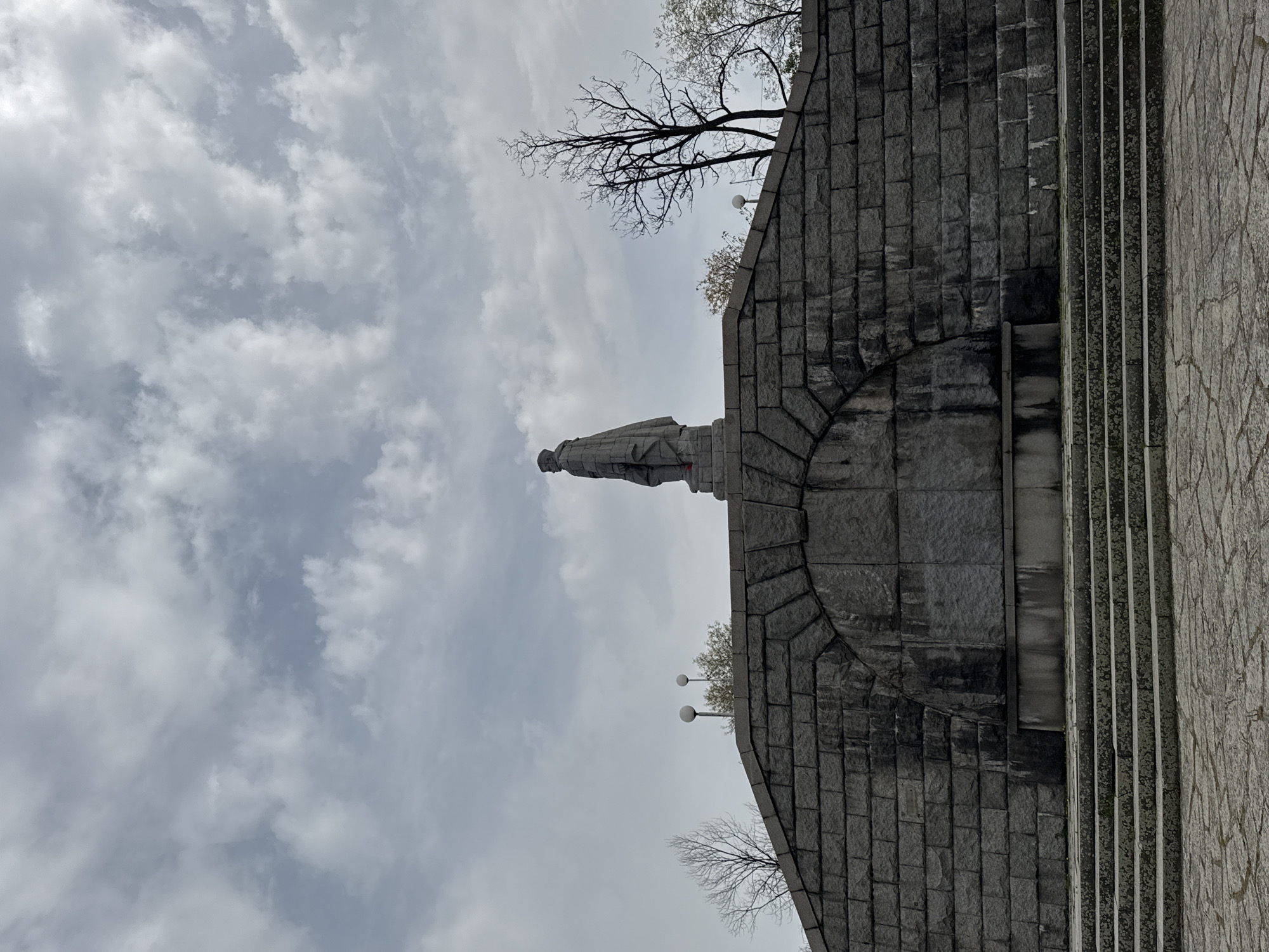 The image shows the Alyosha Monument in Plovdiv, Bulgaria, from a low angle, looking upwards. The monument is a concrete statue of a Soviet soldier, erected atop Bunardzhik Hill. In the background is a cloudy sky with some bare trees. The monument is placed on a stone platform, which consists of a brick base with a decorative arch above. The stairs beside the platform are gray stone.