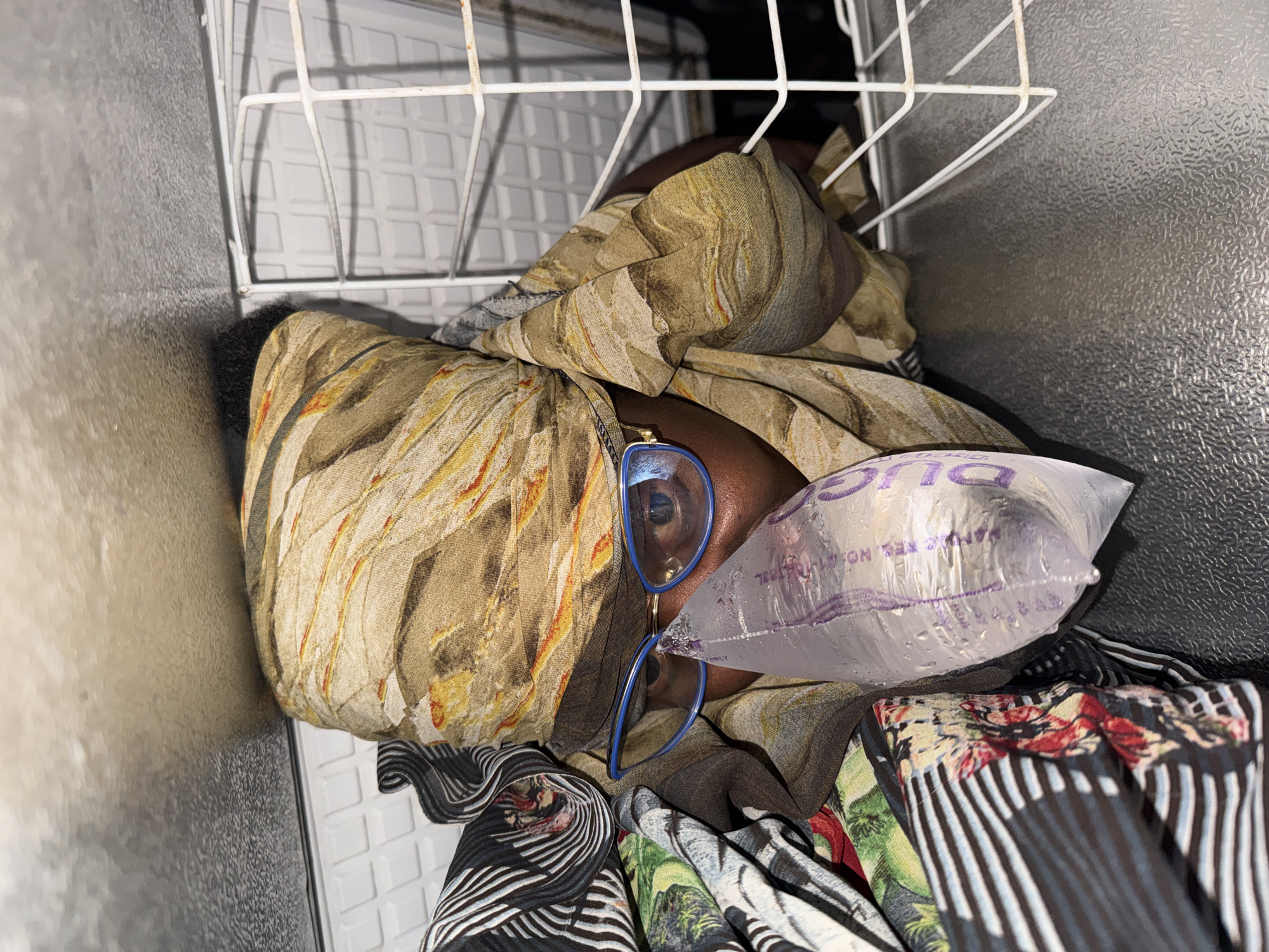 This image shows a person lying down inside what appears to be a freezer. The person is partially covered by patterned fabric. A clear plastic bag, possibly containing ice, is placed near the person's face, and a pair of blue-rimmed glasses are resting on their face, partially obscured by the fabric. The interior of the freezer is visible, with white wire shelving and textured metallic walls. The lighting suggests it is daytime, but no specific time can be determined. The overall impression is one of stillness and a slightly unusual, perhaps uncomfortable, situation. Some text is visible on the plastic bag, appearing to be "MANUF. REG. NO." followed by a number and other unreadable text. The location is identified as Yelwa, Nigeria.