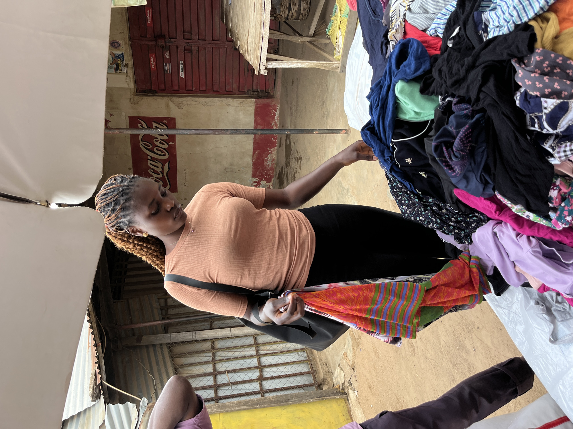 A young woman with brown and black braided hair is shopping for clothing at a market in Gura Top, Nigeria. She wears an orange t-shirt and black pants with a black shoulder bag. She looks down at a pile of used clothing on a white plastic sheet, fingering through the items. Various colors and patterns are visible in the clothing pile, including blue, black, pink, purple, and a striped orange fabric. In the background, there is a building with a red door and a faded Coca-Cola sign. The market seems to be under a corrugated metal roof.