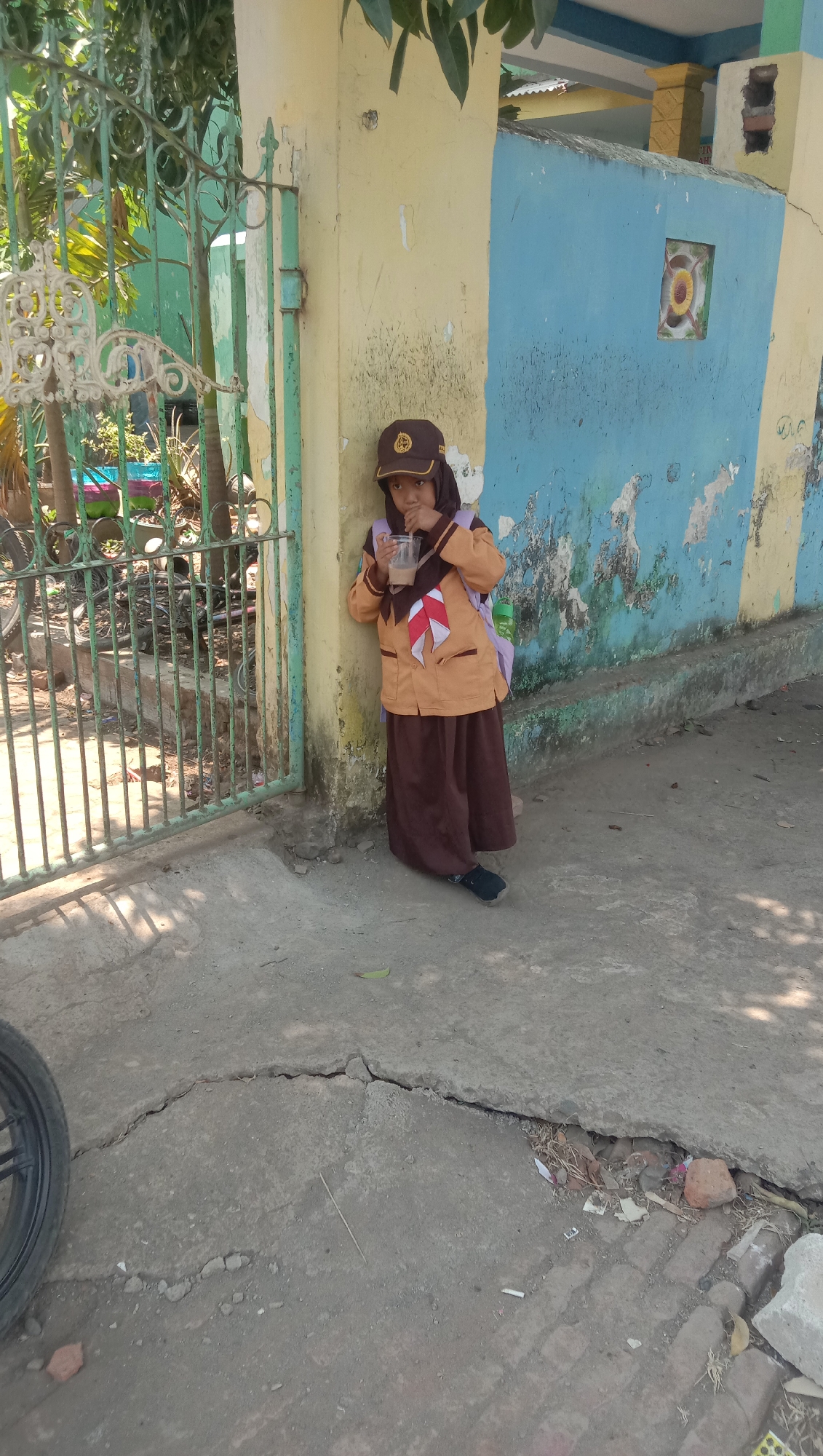 A young Indonesian girl, dressed in a brown and tan scout uniform, stands next to a yellow and blue painted wall, leaning against it with her left shoulder. She is holding a clear plastic cup with a straw in both hands, bringing it to her mouth as if drinking. The girl wears a brown scout hat with a crest and a scarf with red and white elements. A light purple backpack is slung over her right shoulder. The background features a green metal gate, some foliage, and the textured wall of a building. The ground in front of the girl is a cracked concrete path, with some debris and vegetation visible in the cracks. The scene appears to be outdoors, likely in a public area.