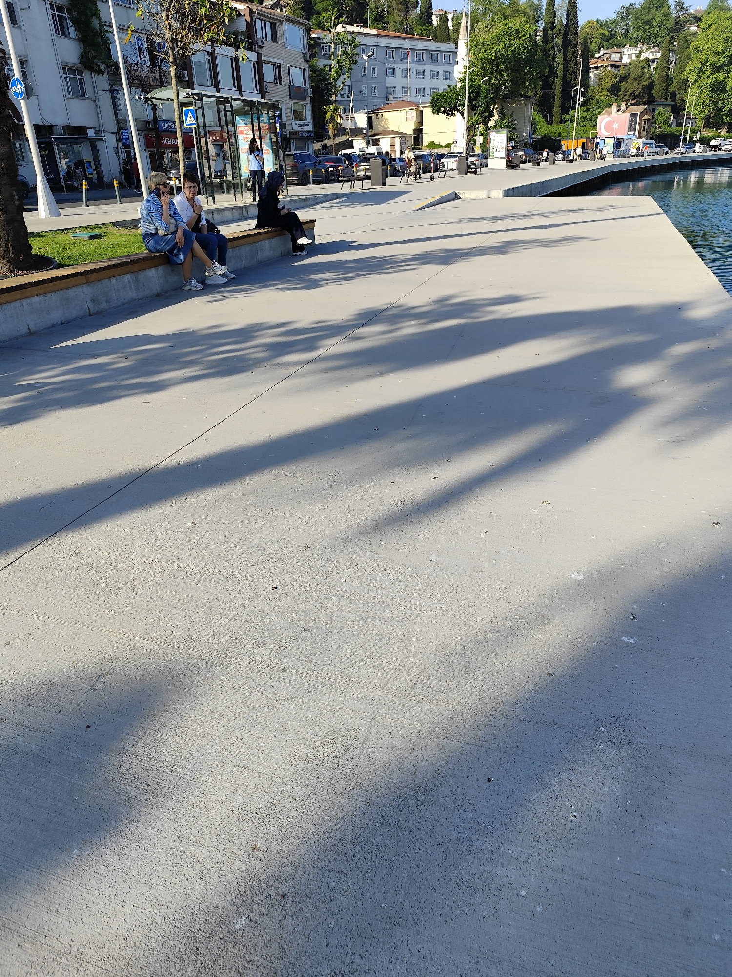 This image depicts a sunny outdoor scene along a waterfront promenade in Sarıyer, Turkey. Long, distinct shadows from trees and overhead structures stretch across the light grey concrete pavement, suggesting either morning or late afternoon.
In the foreground, two women and another individual are seated on a modern wooden bench with a concrete base. The woman on the left, with light-colored hair, is wearing a light top and denim skirt and appears to be speaking on a phone. Beside her, a dark-haired woman in a white top and dark pants is also engaged in conversation. Further down the bench, a person in a dark hoodie sits observing the surroundings.
Behind the bench, a street features parked cars and pedestrians. A glass-paneled bus stop shelter is visible, along with multi-story buildings that line the road. A blue circular sign with a white bicycle symbol is mounted on a pole near the bench. In the background, beyond the buildings, lush green trees cover hilly terrain. On the right, a calm body of water borders the promenade, with a concrete platform extending into it. Further across the water, a building displays a large Turkish flag, featuring a white star and crescent on a red background. Partially visible text on a sign near the bus stop suggests "AKBANK." The overall mood is relaxed, capturing a typical day in an urban waterfront setting.