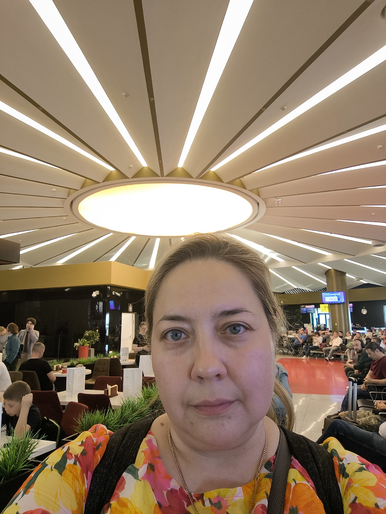A close-up selfie depicts a fair-skinned adult female with light-colored hair and blue eyes, wearing a yellow, orange, and pink floral top with a thin gold-colored necklace and a dark bag strap. Her face occupies the lower foreground, looking directly forward with a neutral expression.
The background reveals a large, modern public interior, specifically an airport or transit lounge in Unknown, Unknown. The dominant feature is a cream-colored ceiling with a central large circular light fixture surrounded by numerous radiating trapezoidal panels. Alternate panels contain recessed linear strip lighting, all brightly illuminated. Thin dark lines separate these panels, with some golden-colored structural elements visible near the central light. Multiple small white smoke detectors are mounted on the ceiling panels.
To the left of the subject, a cafe-like area features dark walls topped with a golden band. White tables with dark chairs are arranged, interspersed with green potted plants. Several individuals are present; a young boy in a black shirt is seated at a table, resting his head on his hand.
To the right, a large, light-colored cylindrical column stands. Beyond it, a spacious waiting area is visible with multiple individuals seated in rows of chairs. A digital display screen showing "09" is mounted on a far wall. The floor in the background transitions from a reddish-orange section to darker areas. The environment is well-lit and appears to be a busy public space.