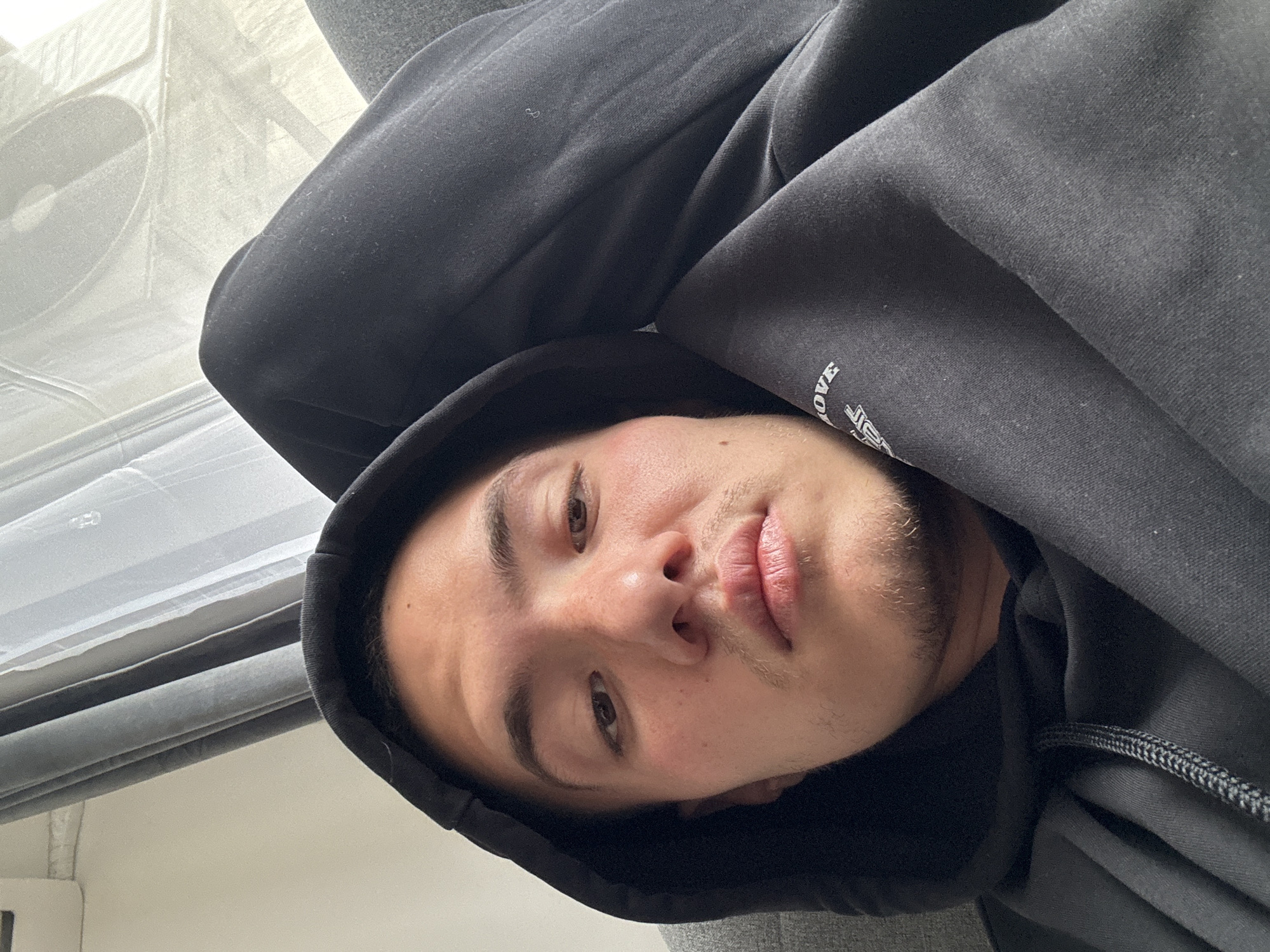 A selfie shot from a low angle depicts a male with light brown skin, brown eyes, and short dark hair wearing a black hooded sweatshirt. The hood is pulled up, framing his face. He has a stubble beard and mustache. The sweatshirt's material appears to be thick and soft. To the left of the frame, a curtain is visible. It is unclear if the image was taken in the city of Varna, Bulgaria.