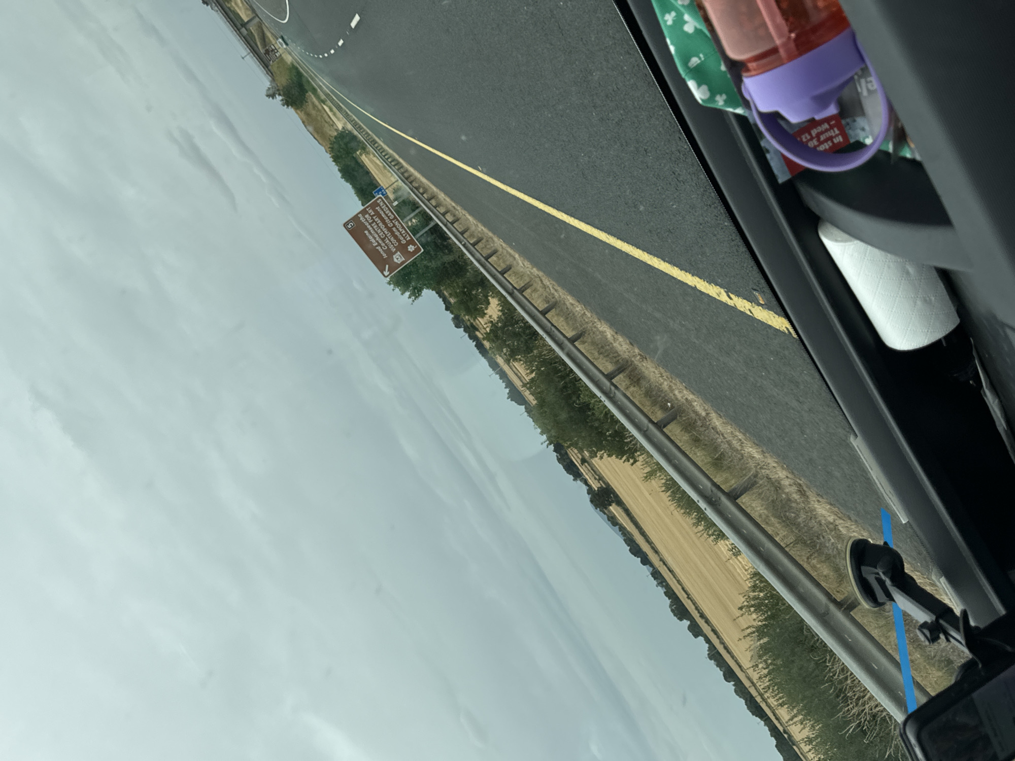The image is a roadside view from inside a vehicle traveling on a highway in the city of Moyle, Ireland. The sky is overcast and cloudy. On the right side of the road, a brown directional sign is visible, indicating points of interest such as "Ionad Ealaine," "Cothabhalaimhth," "INSUL CENTRE FOR CONTEMPORARY ART," "Cathair Gheanainnt," and "Altachont Gardens." The road has a yellow line dividing the lanes. The surrounding landscape features fields of golden crops and green trees. In the foreground, inside the vehicle, a cell phone mount is attached to the dashboard, and a water bottle and a roll of toilet paper are visible.