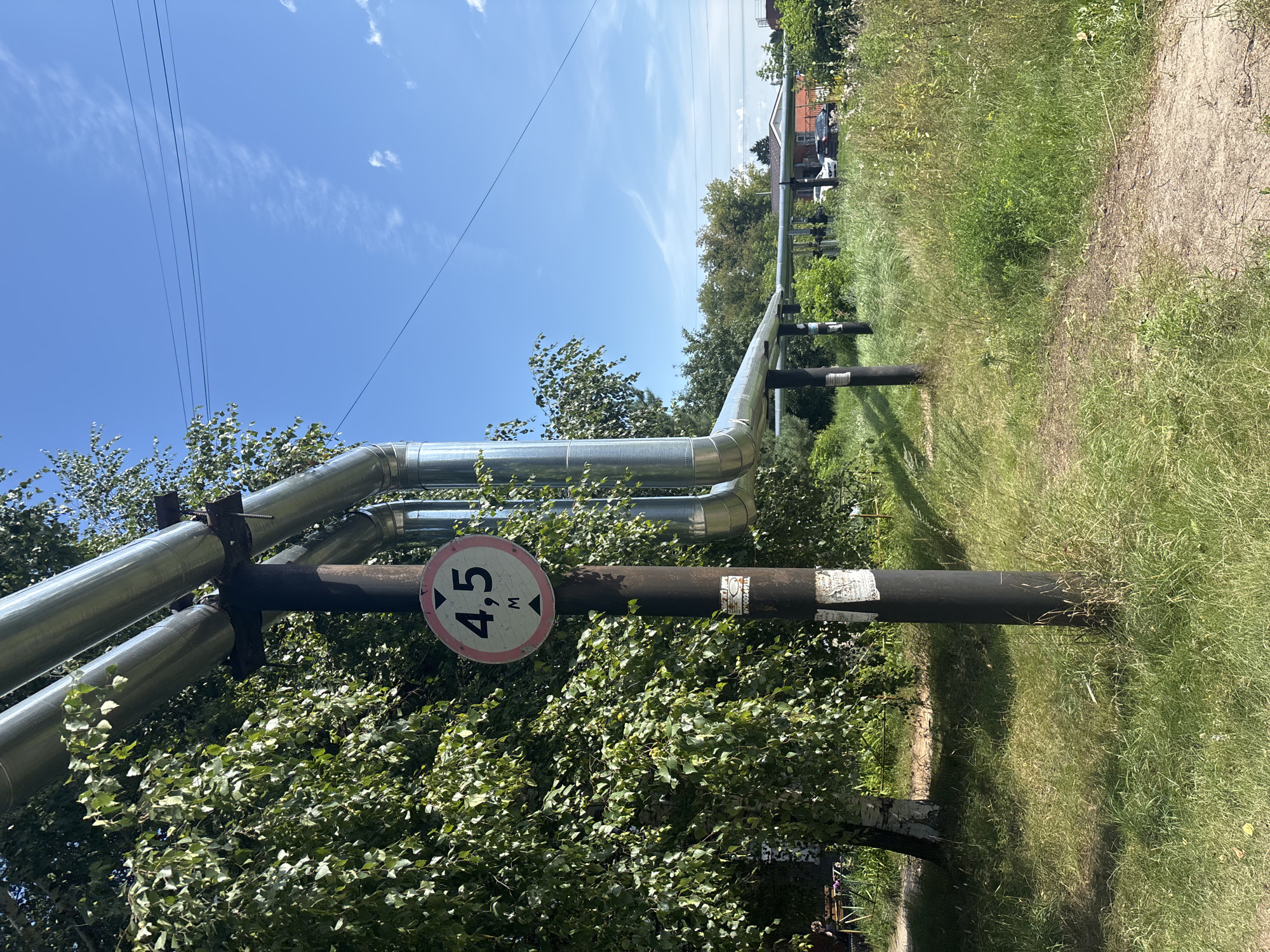 The image shows a section of overhead pipes, likely for district heating, running parallel to a dirt path. A round sign with pink and white borders is attached to one of the support poles, displaying "4,5 M" with arrows pointing upwards and downwards, indicating a height restriction of 4.5 meters. The pipes are silver and supported by dark metal poles. To the left, there are green birch trees with dense foliage. The ground is covered in grass and some wildflowers. In the background, there are buildings and cars visible, suggesting a suburban or residential area. The sky is clear blue with a few wispy clouds and overhead power lines. There are no people clearly visible in the immediate vicinity of the pipes and sign. The scene appears to be in Kstovo, Russia, given the presence of district heating infrastructure common in many Russian cities and the visible style of the houses in the background.