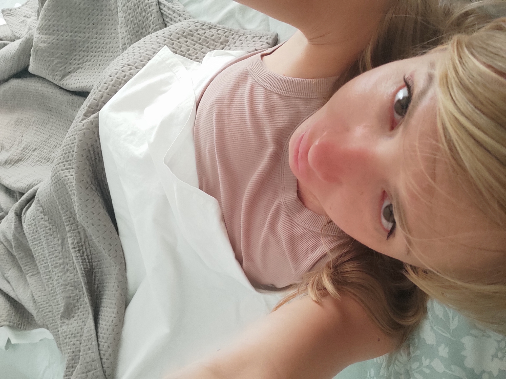 The image shows a close-up, upside-down selfie of a blonde woman lying in bed. She is wearing a pink ribbed tank top. Her arms are raised above her head, and she is covered by white sheets and a grey textured blanket. The bed appears to be in a bedroom with light blue floral patterned sheets visible at the bottom of the frame. The overall impression is of a relaxed, intimate setting. The location in Blanes, Spain is not discernible from the image.