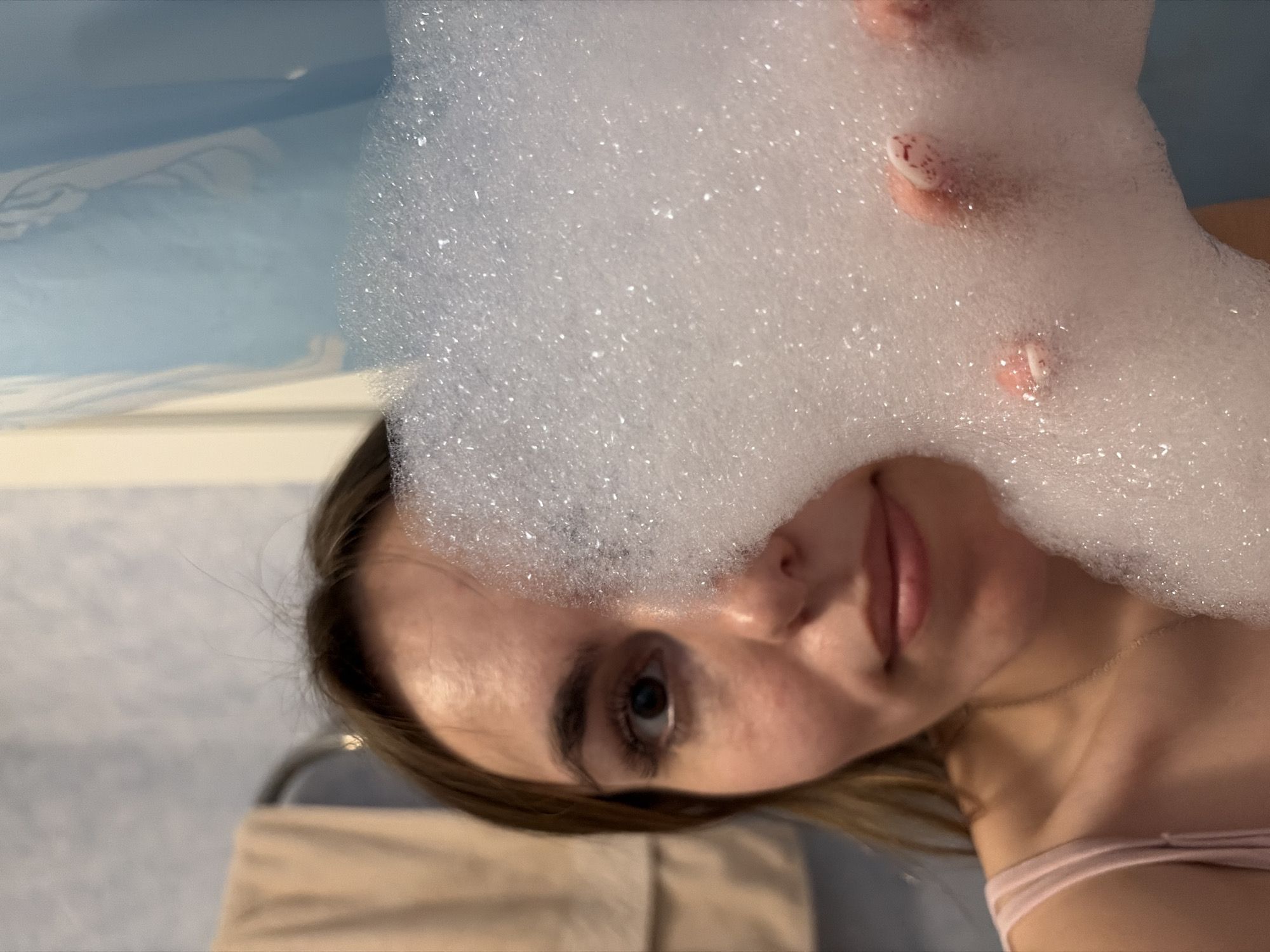 A person with light skin and brown hair is partially obscured by white soap bubbles. The bubbles are also covering the person's hands. The person is in a bathtub with blue edges. A white towel rack is visible in the background. The location in Izhevsk, Russia is not discernible.