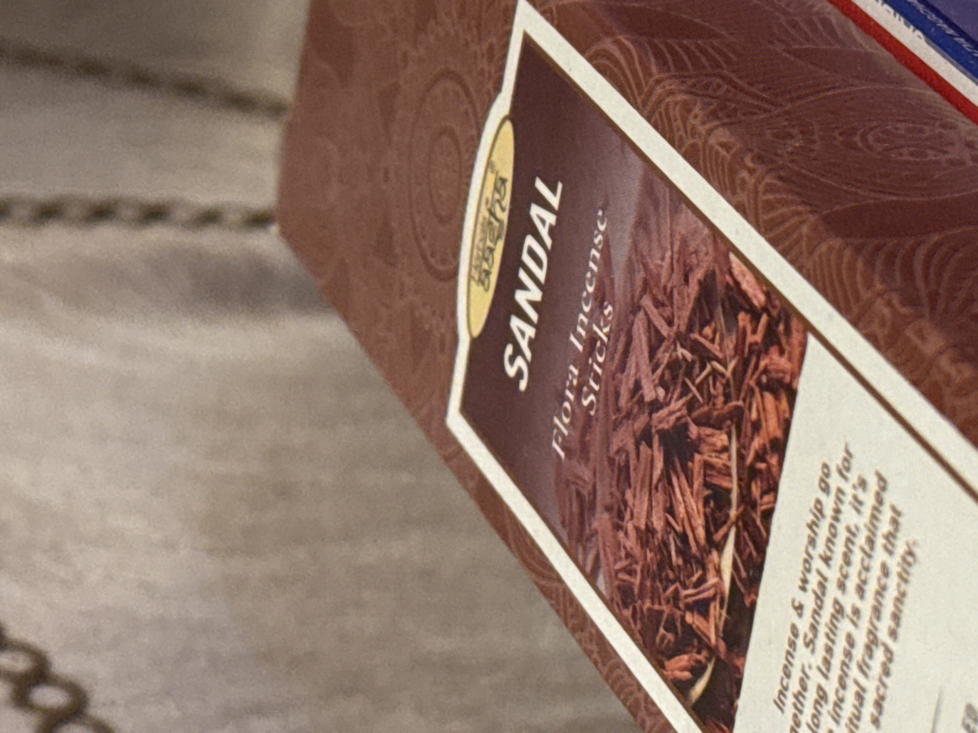 The image shows a close-up of a box of "Satya Sandal Flora Incense Sticks". The text on the box includes the words "Sandal" and "Flora Incense Sticks". A description of the incense is also visible, stating that it is known for its long-lasting scent and is acclaimed for its spiritual fragrance and sacred sanctity. The background is a blurred pattern, possibly part of the box's design or the surface it's sitting on. There is no indication of the location being Kladovitsy, Russia in the image.