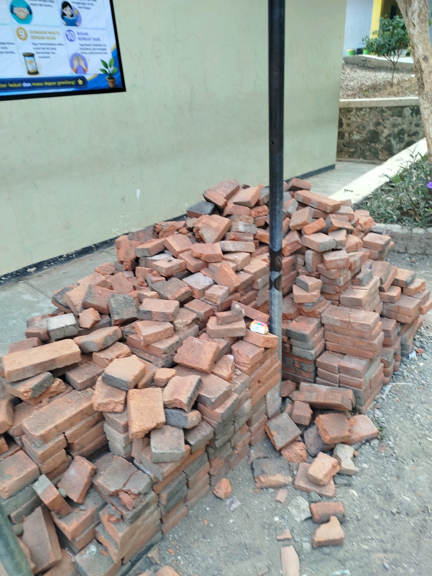 A pile of used red bricks is situated on a gray, gravelly ground in front of a light green wall. The bricks are of various sizes and conditions, with some appearing broken and weathered. A dark, vertical pole stands among the bricks. In the background, a tree trunk is visible to the right, and beyond that, a textured stone wall and a small green tree. A blue and yellow poster with text and illustrations is mounted on the wall on the left. The scene appears to be outdoors, likely in a construction or storage area. The presence of the poster suggests an educational or community setting. The location is specified as Kedungwaru, Indonesia.
