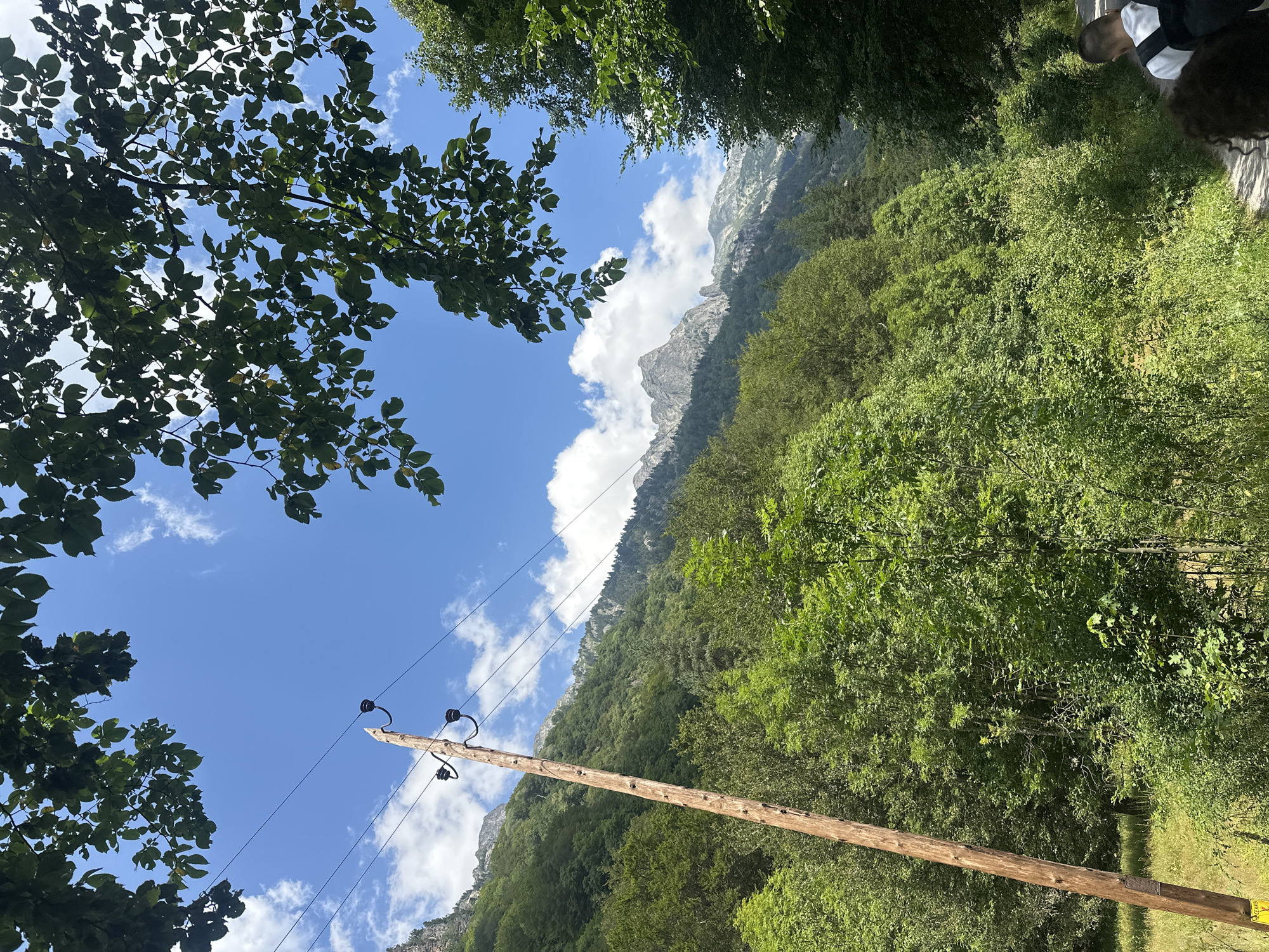 The image depicts a rural scene with a prominent wooden telephone pole on the left, extending upwards against a bright blue sky with scattered white clouds. The pole is adorned with insulators and power lines. The surrounding environment is lush and green, characterized by dense vegetation, including trees and bushes, suggesting a natural, possibly mountainous landscape. In the background, rugged, tree-covered mountains rise against the sky. The lower right portion of the image shows a dirt path, with the backs of two individuals visible, suggesting they are walking along the path. The overall impression is one of a serene, outdoor setting. There is no information about this being in Rilski manastir, Bulgaria.