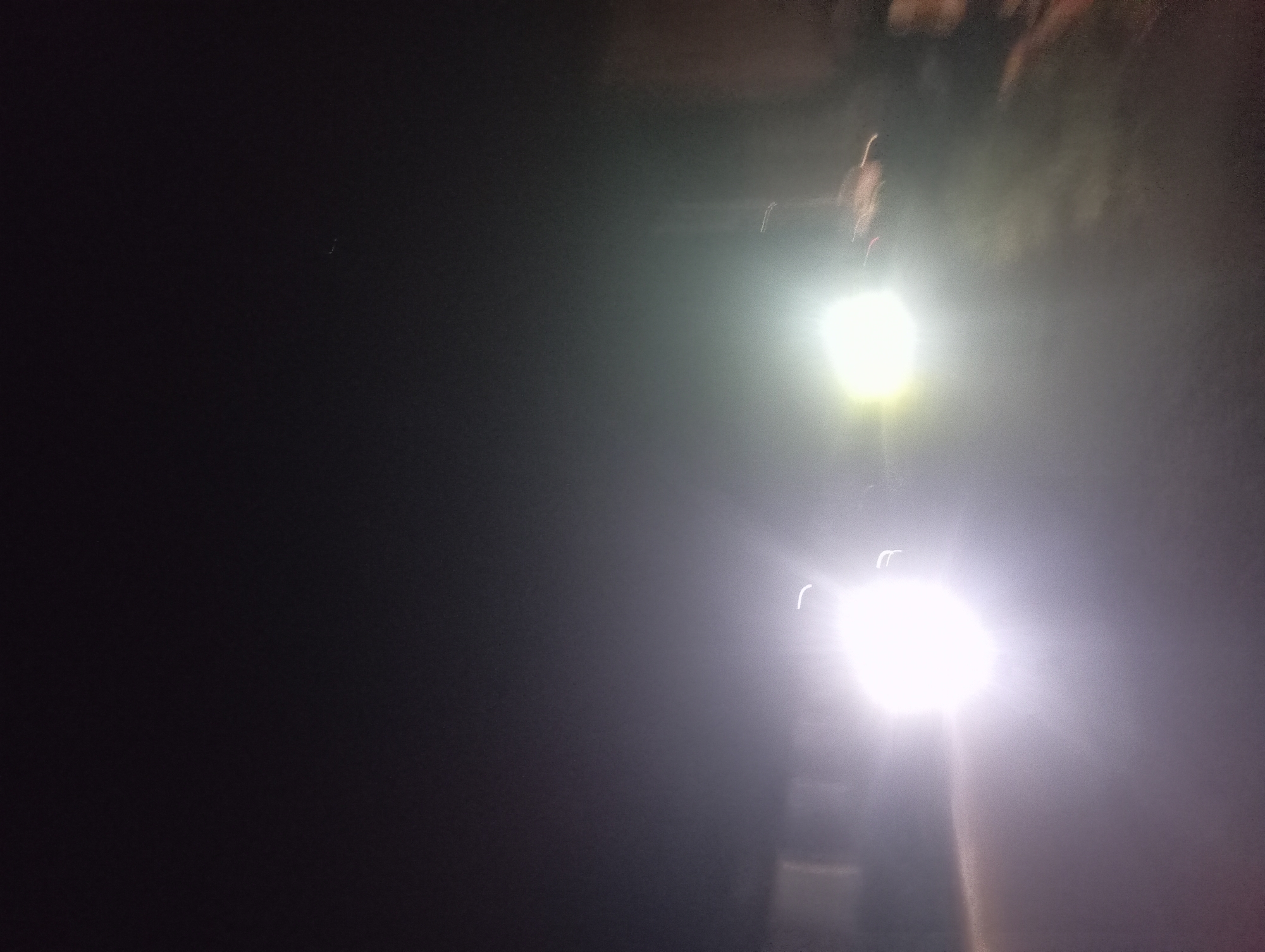 The image shows two bright streetlights illuminating a dark scene in Nkpor, Nigeria. The left side of the image is predominantly black, transitioning to lighter shades towards the right where the streetlights are located. The streetlights are vertically aligned, and their beams create a halo effect in the surrounding air. The upper portion of the image suggests the presence of trees or other overhead structures. The overall scene is dark and obscured by the light of the streetlights.