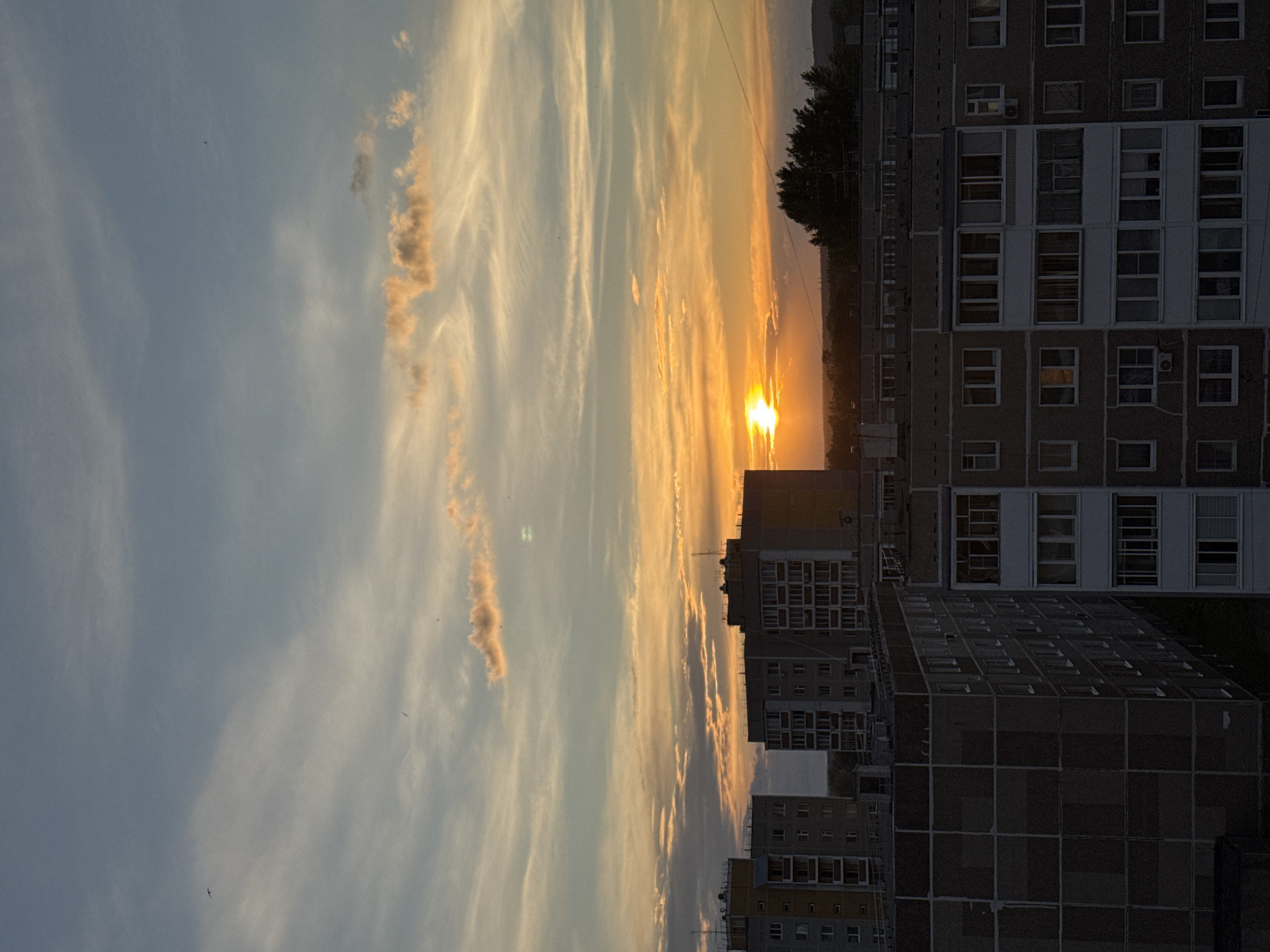 This is a medium-shot, vertical, exterior photograph taken during sunset in the city of Chaikovsky, Russia. The image captures a residential cityscape with apartment buildings and a vibrant sky filled with clouds.
In the foreground, the tops of several large, multi-story apartment buildings are visible. These buildings appear to be constructed of concrete and feature numerous windows and balconies. Their facades are textured and somewhat weathered. Some buildings are darker in color, while others have lighter-colored sections.
The middle ground is occupied by more apartment buildings, some of which are taller and appear to be further away. These structures extend across the frame, suggesting a dense urban environment. Trees are visible in the distance, silhouetted against the sunset.
The background is dominated by the sky, which is a spectacle of sunset colors. The sun is a bright, orange orb, partially obscured by clouds, casting a warm glow across the scene. Wispy clouds are scattered across the sky, colored in shades of orange, yellow, pink, and blue. The overall atmosphere is serene and picturesque, characteristic of a sunset over a city.
There are no visible people in the photograph. The focus is on the architecture and the natural phenomenon of the sunset. The lighting suggests it is late evening. The image appears to be taken from a high vantage point, looking down on the city.