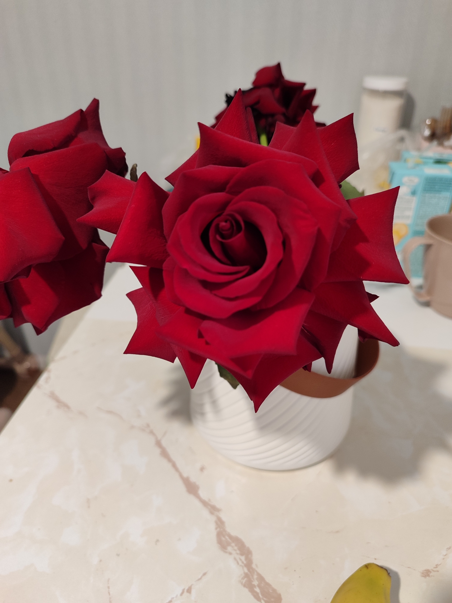 The image presents an indoor still life composition featuring three vibrant red roses arranged in a white, horizontally ribbed planter with a brown top rim. The central rose is in sharp focus, showcasing its intricate, spiraled petals and rich, deep red color. Another rose is visible to the left, slightly out of focus, while a third is partially obscured behind the main rose.
The planter rests on a light-colored surface, likely a countertop or table, characterized by a subtle marble-like pattern with beige and brown veining. In the blurred background, several domestic items are visible. These include a white-lidded jar or container, a light blue carton (possibly for beverages), and a beige or light brown mug situated to the right of the floral arrangement. A yellow object, appearing to be a banana, is also visible at the bottom right corner of the frame.
The lighting suggests an indoor setting, likely artificial, without indicating a specific time of day. No human presence or activity is depicted. The scene is static, focusing on the aesthetic display of the roses. There is no visible text in the image. While the location context is provided as Izhevsk, Russia, no specific visual cues within the image confirm this geographical detail.