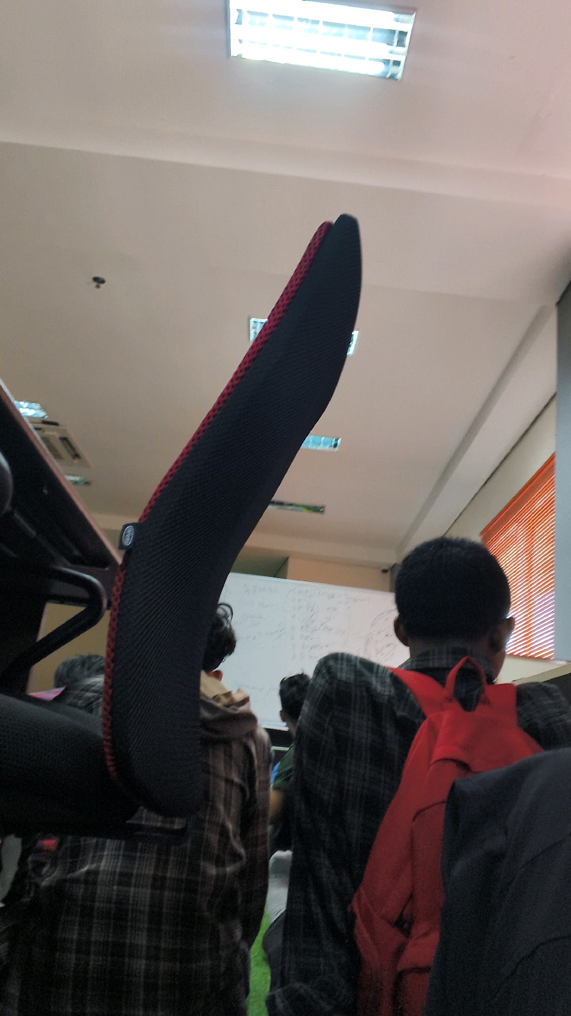 The image depicts an indoor scene, likely a classroom or lecture hall, with fluorescent lighting overhead. In the foreground, the curved backrest of a black and red mesh chair is prominently displayed, extending diagonally upwards. To the right, the back of a person wearing a plaid shirt and a red backpack is visible. Further back, another individual in a plaid shirt can be seen. A whiteboard with writing is visible in the background, and a window with orange blinds is to the far right. The presence of multiple people suggests a gathering or educational setting. The image quality is moderate, with some blur and graininess, particularly in the background. The specific location in Lohbener, Indonesia, cannot be determined from the visual information alone.