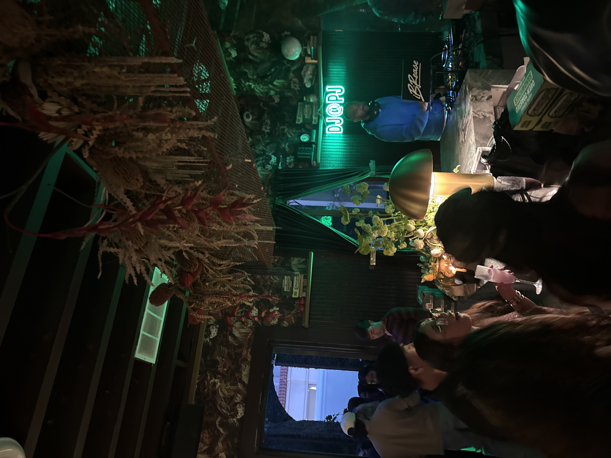 This vertical image captures the vibrant, dimly lit interior of a bar or lounge in Los Angeles, United States of America, likely during the night. The scene is illuminated by green neon lights and various accent lamps, creating a lively atmosphere.
In the background, a DJ stands behind a booth, wearing a blue long-sleeved shirt and headphones. Above the DJ, a prominent green neon sign glows, spelling out "DJ@PJ". To the right of the DJ booth, a black panel features the text "iB Rease". The booth itself holds DJ equipment, including what appears to be turntables or mixers. Behind the DJ, shelves display decorative items, including a clock and what looks like small globes or spheres.
The midground and foreground are occupied by several patrons. On the right, a woman with long, dark hair is clearly visible, holding a stemmed glass containing a pink drink. She appears to be socializing with others around her. People in the immediate foreground are partially obscured, adding to the sense of a crowded venue. A distinct golden, mushroom-shaped lamp provides warm light on a table in front of the crowd. Also on the table is a cardboard box with the text "OVEN-TOASTED SANDWICHES" printed on it.
The decor includes abundant dried floral arrangements and intricate leafy structures hanging from the ceiling and adorning the walls, giving the space an eclectic, natural, or possibly bohemian aesthetic. Behind the crowd, a window offers a glimpse of the outside, showing a building with a blueish artificial light, confirming the nighttime setting. The overall mood suggests a relaxed social gathering enjoying a DJ set.