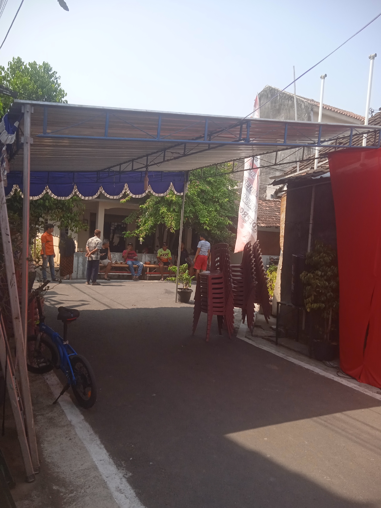In Surakarta, Indonesia, an outdoor event is underway beneath a temporary canopy structure. The canopy, made of corrugated metal and supported by a blue metal frame, is set up over a paved area. A blue and white banner hangs from the canopy. In front of a building, several people are gathered, some sitting on benches and others standing. A stack of red plastic chairs is visible to the right, and a blue bicycle is parked to the left. The scene suggests a public gathering or a temporary outdoor setup, possibly for a local event or celebration in the city.