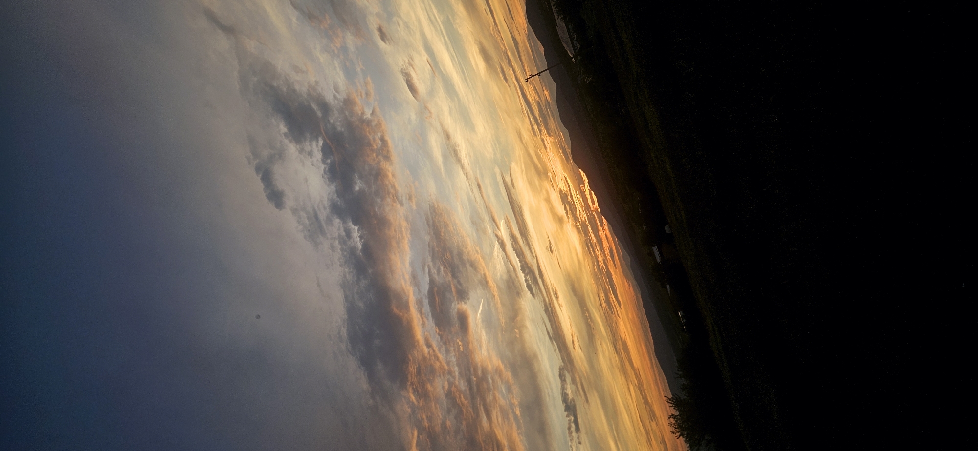 The image captures a dramatic sunset over a rural landscape, likely Momin Prohod, Bulgaria. The sky is ablaze with hues of orange, yellow, and pink, painted across a canvas of deepening blue and gray clouds. Streaks of lighter clouds, possibly contrails, crisscross the scene. In the distance, rolling hills and mountains are silhouetted against the vibrant sky. The foreground is dominated by dark, silhouetted foliage, suggesting a hilly terrain covered in grass and low-lying vegetation. A solitary utility pole stands in the mid-ground on the right, its presence stark against the natural beauty of the sunset. The overall impression is one of a serene, yet intensely colorful, end to the day in a tranquil, undeveloped area.