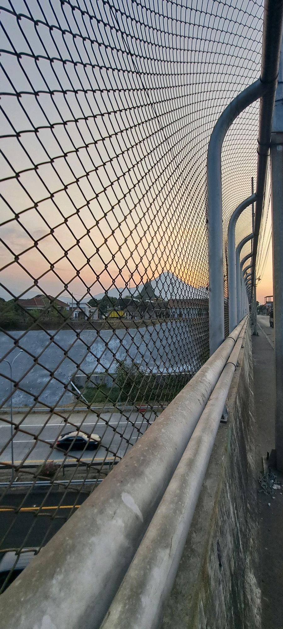 This image depicts a view from a bridge or elevated walkway in Randupitu, Indonesia. In the foreground, a metal chain-link fence is prominent, with thick metal pipes running along the side of the walkway. The fence offers a view of the scene below and beyond.
Below the fence, a road is visible with a single dark-colored car in motion. The road appears to be a multi-lane highway or significant thoroughfare. Adjacent to the road is a body of water, possibly a river or canal, with a concrete retaining wall bordering it.
In the middle ground, a small settlement or village is visible, with houses and buildings, including structures with distinctive Islamic architectural elements such as domes and minarets. A large, dark, mountainous silhouette dominates the background, suggesting the presence of a volcano or significant mountain range.
The sky displays the warm hues of either sunrise or sunset, with soft oranges, yellows, and pinks blending into a pale blue. The overall impression is a serene, dusky scene in a semi-urban or rural Indonesian setting.