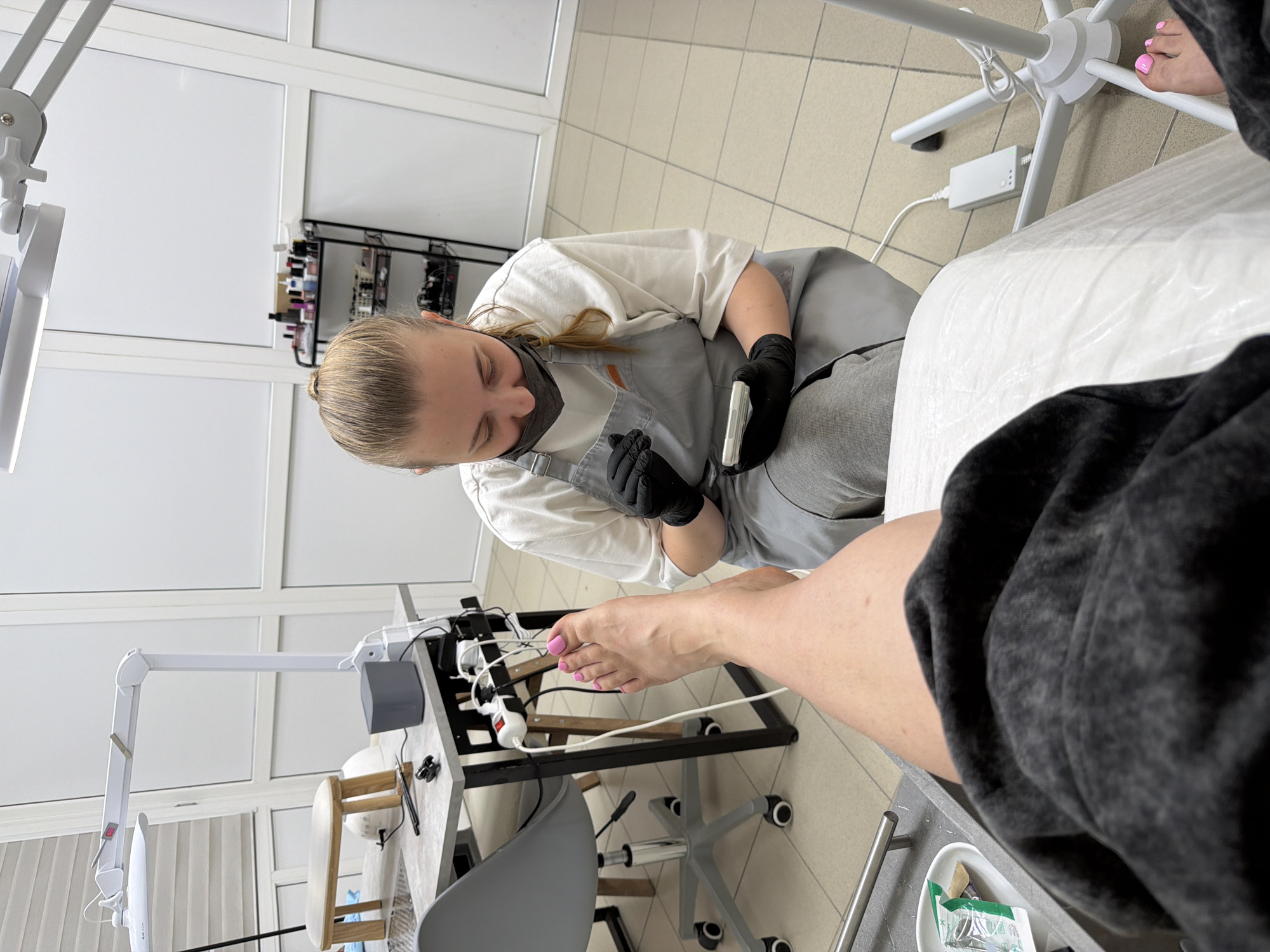 In Leningradskaya, Russia, a woman is seated on the floor, wearing black gloves and a grey apron over a white t-shirt and grey sweatpants. She is holding a white electronic device and looking down at it. In front of her is a person's foot with bright pink toenails, resting on a white surface. The environment appears to be a nail salon, with equipment such as a lamp, a table with tools, and a rolling chair visible. In the background, there is a shelf unit filled with bottles and a tiled floor and wall.