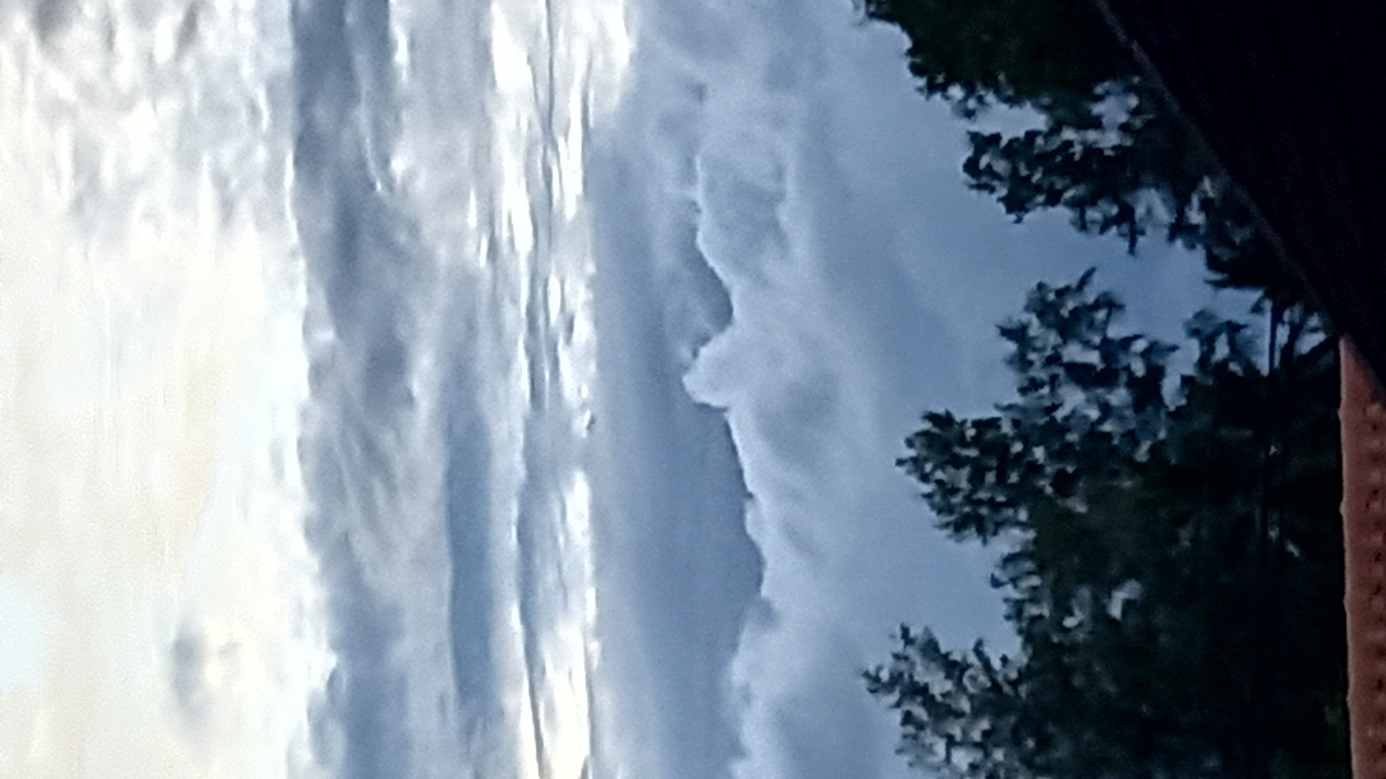 The image is a vertical shot of a cloudy sky over trees. The sky has layers of clouds, with brighter white clouds near the top and darker, grayer clouds lower down. In the foreground, silhouetted dark green trees are visible, with their foliage creating an irregular pattern against the sky. The bottom of the image shows a sliver of a reddish-brown tiled roof. The scene appears to be outdoors, possibly in a residential area given the presence of a roof. The location of Ko Pha-ngan District, Thailand, is not discernible from the image content itself, as there are no specific landmarks or identifying features.