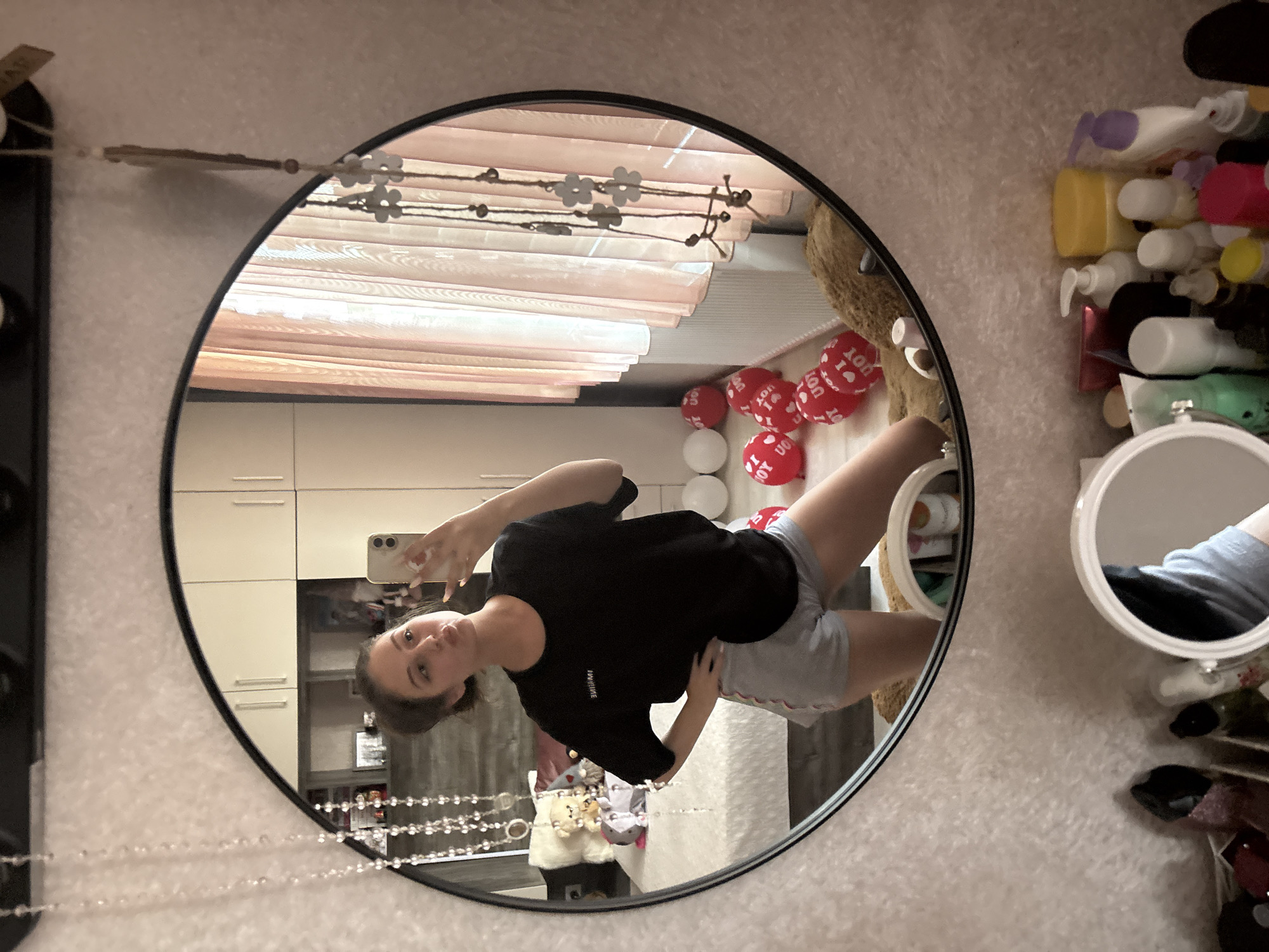 The image shows a young woman taking a mirror selfie in what appears to be a bedroom or dressing area. She is wearing a black t-shirt and grey shorts, with her hair pulled back. She is holding a smartphone in her right hand and posing with her left hand on her hip. In the background, reflected in the mirror, is a bed with several red and white balloons, a teddy bear, and a closet with white doors. To the right of the mirror, pink curtains are visible. Below the large mirror, on a surface that looks like a dresser or vanity, are various bottles and containers of toiletries and cosmetics. The overall scene is brightly lit, suggesting it is daytime. The location is identified as Bulgarovo, Bulgaria.