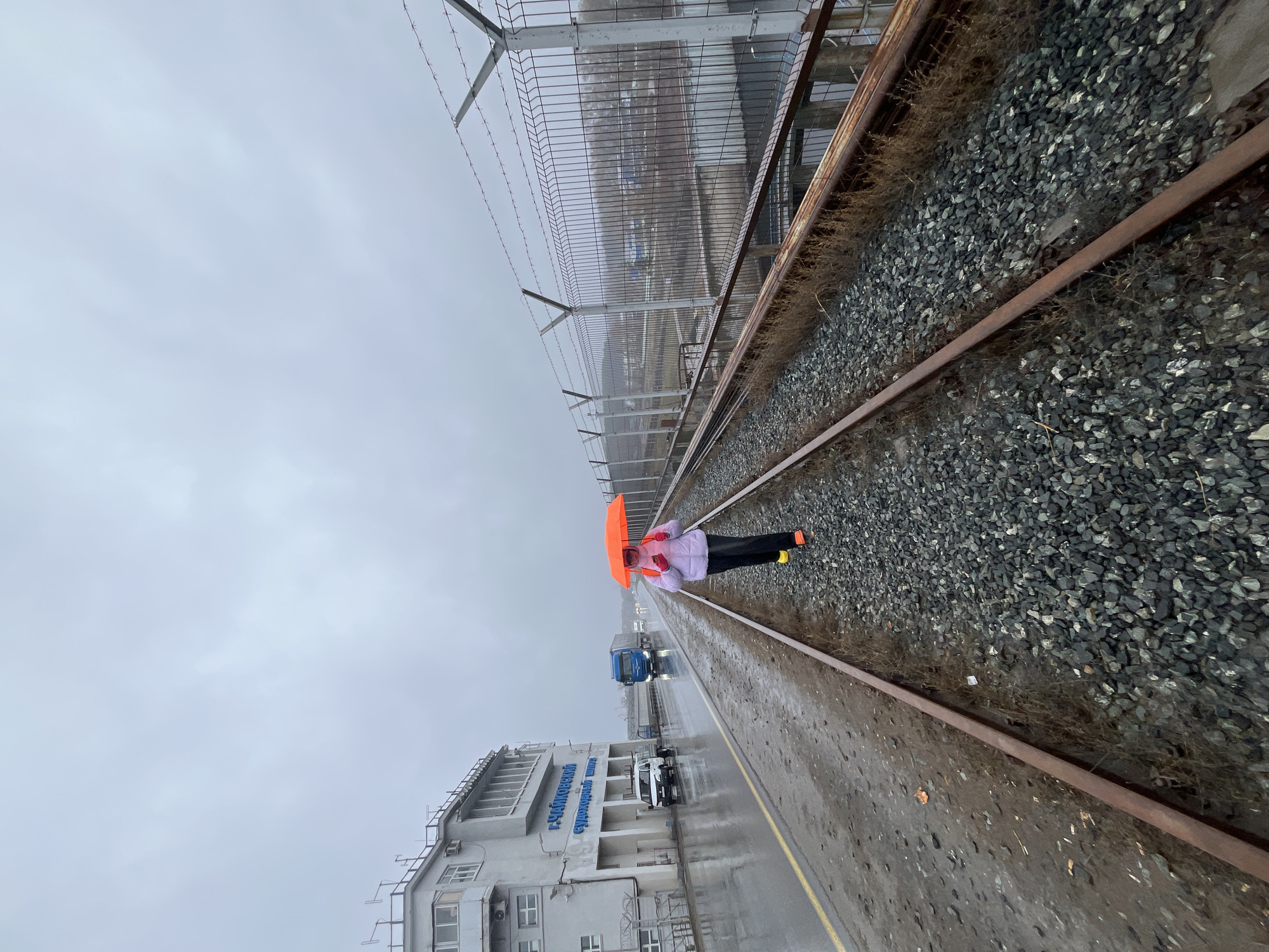 This image, presented in a sideways orientation, depicts an outdoor industrial scene in Chaikovsky, Russia, under an overcast, grey sky, suggesting a cool, damp day. The ground, including a paved road and railway tracks, appears wet from recent rain or ongoing precipitation.
In the middle ground, a person stands on two parallel railway tracks, holding a bright orange open umbrella above their head. The individual is dressed in a light-colored jacket, dark pants, and features bright orange accents near their feet. The railway tracks are rusty and embedded in dark grey gravel ballast, with sparse dried vegetation growing alongside them.
To the right of the tracks (when viewed upright), a tall, robust metal fence topped with barbed wire extends into the distance, separating the tracks from an unseen area. To the left of the tracks, a dark, wet paved road or path with a yellow line runs parallel. Adjacent to this road is a multi-story industrial building, light in color, featuring numerous windows, external ladders, and air conditioning units. Prominently displayed on its facade is blue Cyrillic text that reads "ПАО «Куйбышевазот»" (PAO Kuybyshevazot), indicating a chemical or industrial enterprise. A blue semi-truck and a white car are visible on the wet paved surface near the building.
The scene conveys a functional, industrial atmosphere, with the person on the tracks possibly observing, waiting, or posing amidst the bleak weather conditions.