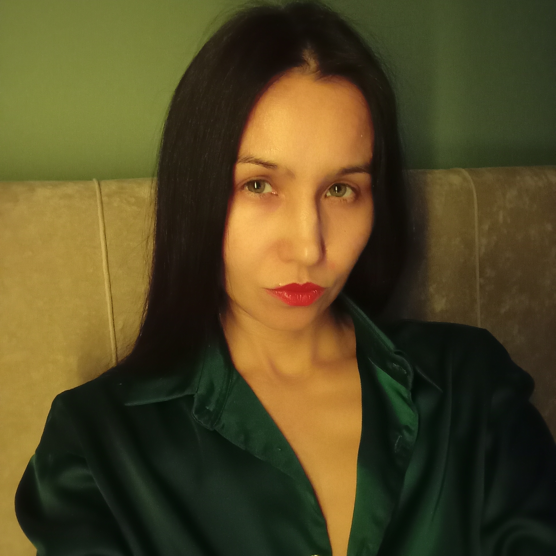 A woman is seated indoors, likely against a headboard or similar cushioned structure. The background is a greenish hue, possibly a wall color. The woman has dark, straight hair, fair skin, and is wearing bright red lipstick. She is wearing a green, silken, collared shirt or blouse that is partially open. The location is not discernible as being in Pervomaysky, Russia from visual information alone.