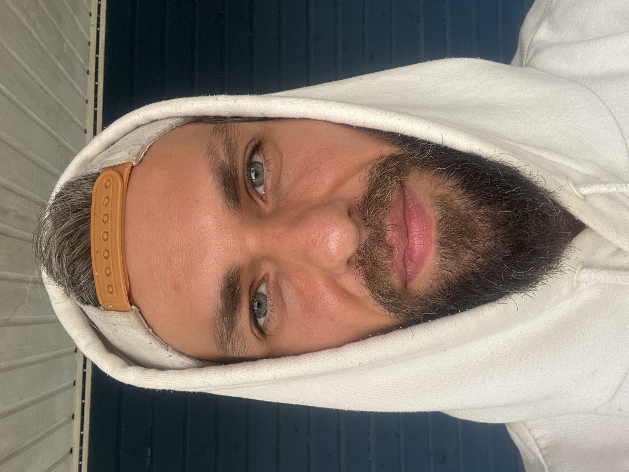The image is a close-up, head-on portrait of a man wearing a white hoodie and a tan baseball cap. He has blue eyes, a full beard, and short, graying hair. The background appears to be a dark blue, horizontally slatted wall, possibly the side of a building. The lighting suggests an outdoor setting, possibly during the day. There is no indication of the city of Kuti, Russia in the image or its description.