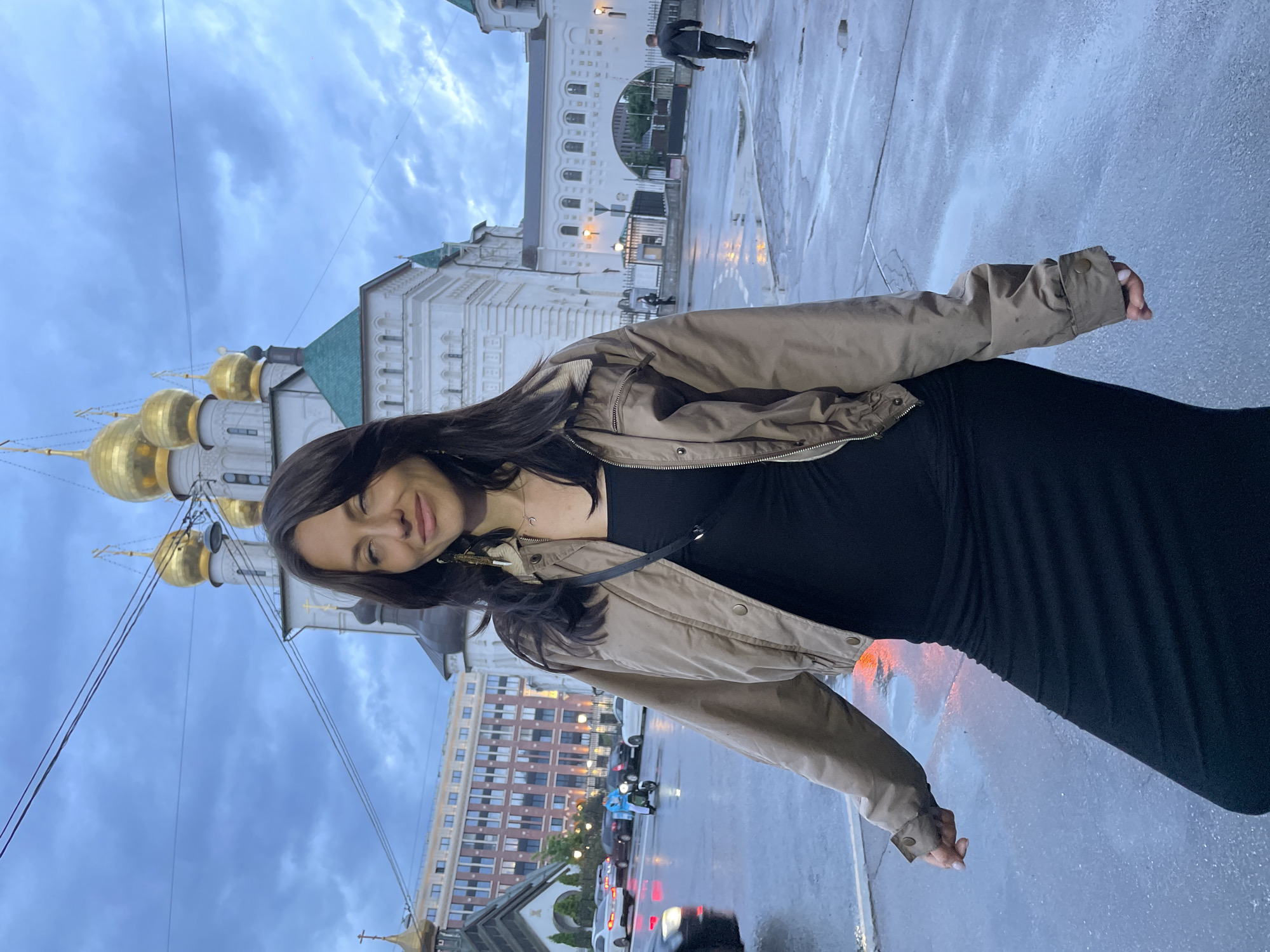A young woman with dark hair and light skin stands in the foreground of the image. She is wearing a beige bomber jacket over a black dress. Her right arm is bent at the elbow, and her left hand is slightly extended. She is looking at the camera with a slight smile and her eyes are partially closed.
In the background, a large white building with gold domes and green roofs is visible. The sky is overcast and appears to be raining, as the ground is wet and reflective. Several cars and a person on a scooter are present in the street. The woman appears to be standing on a wet pavement in front of a building that resembles a cathedral or church. The architecture and the presence of multiple onion domes suggest a location within the historic center of Saint Petersburg, Russia. The woman's attire suggests a casual outing, perhaps exploring the city despite the weather. The overall atmosphere is subdued due to the cloudy skies and wet conditions.