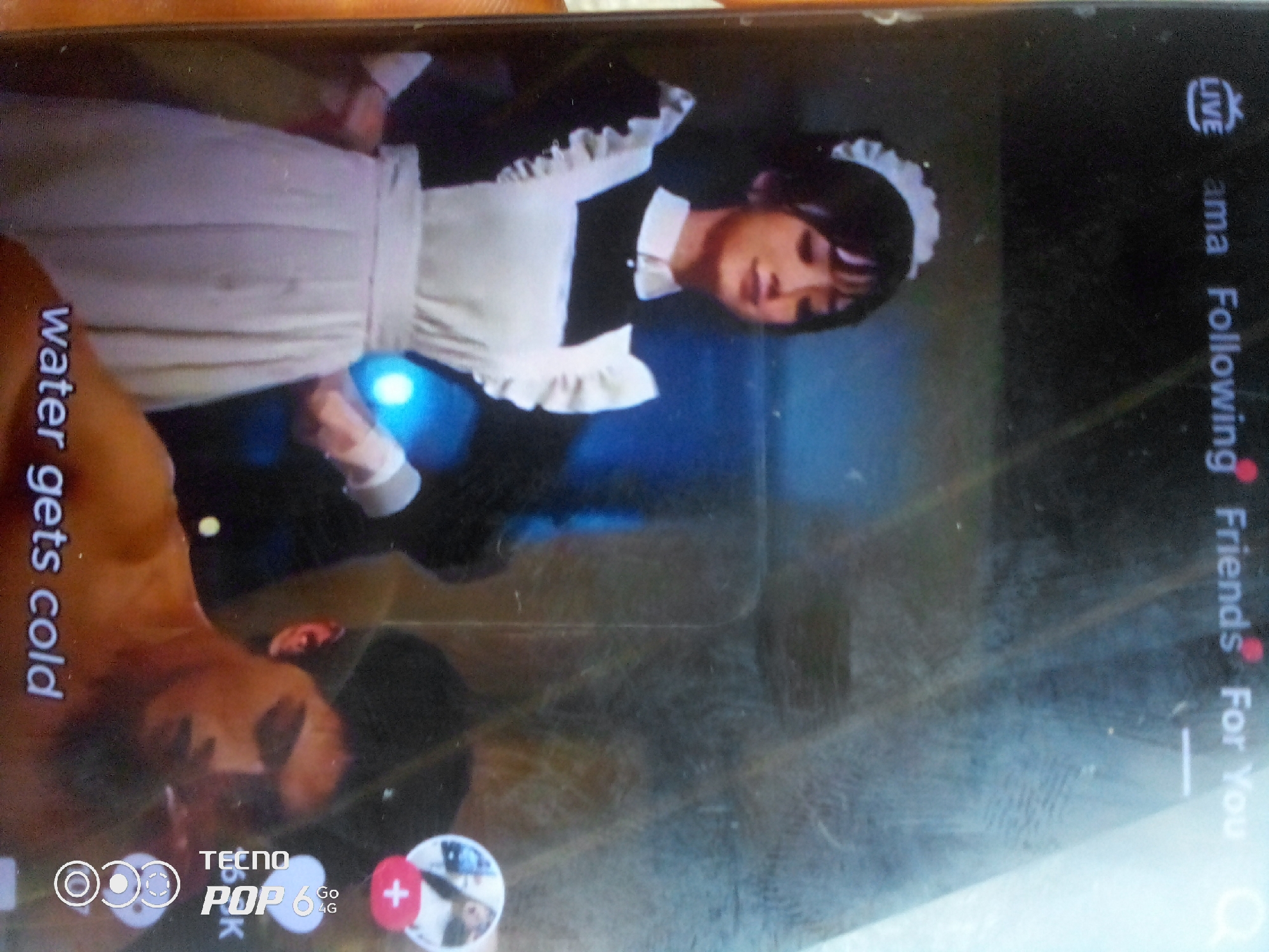 The image displays a close-up of a smartphone screen, viewed from an angle. The screen shows a video or image of a person dressed in a maid outfit, with a black dress and white apron with ruffles. The person's face is partially visible, with dark hair. Text overlays on the screen include "water gets cold" vertically on the left, and "LIVE", "ama", "Following", "Friends", and "For You" vertically on the right. A portion of another person's arm and chest is visible in the lower left corner of the frame, with skin tone appearing brown. The bottom of the screen shows branding for "TECNO POP 6 4G" with icons and a red plus sign. The background of the phone screen appears to be a dark, possibly blueish, setting. The environment outside the phone is not visible. The image is a photograph of the smartphone screen, and thus, the location within Howayi, Nigeria, is not determinable from the provided image.