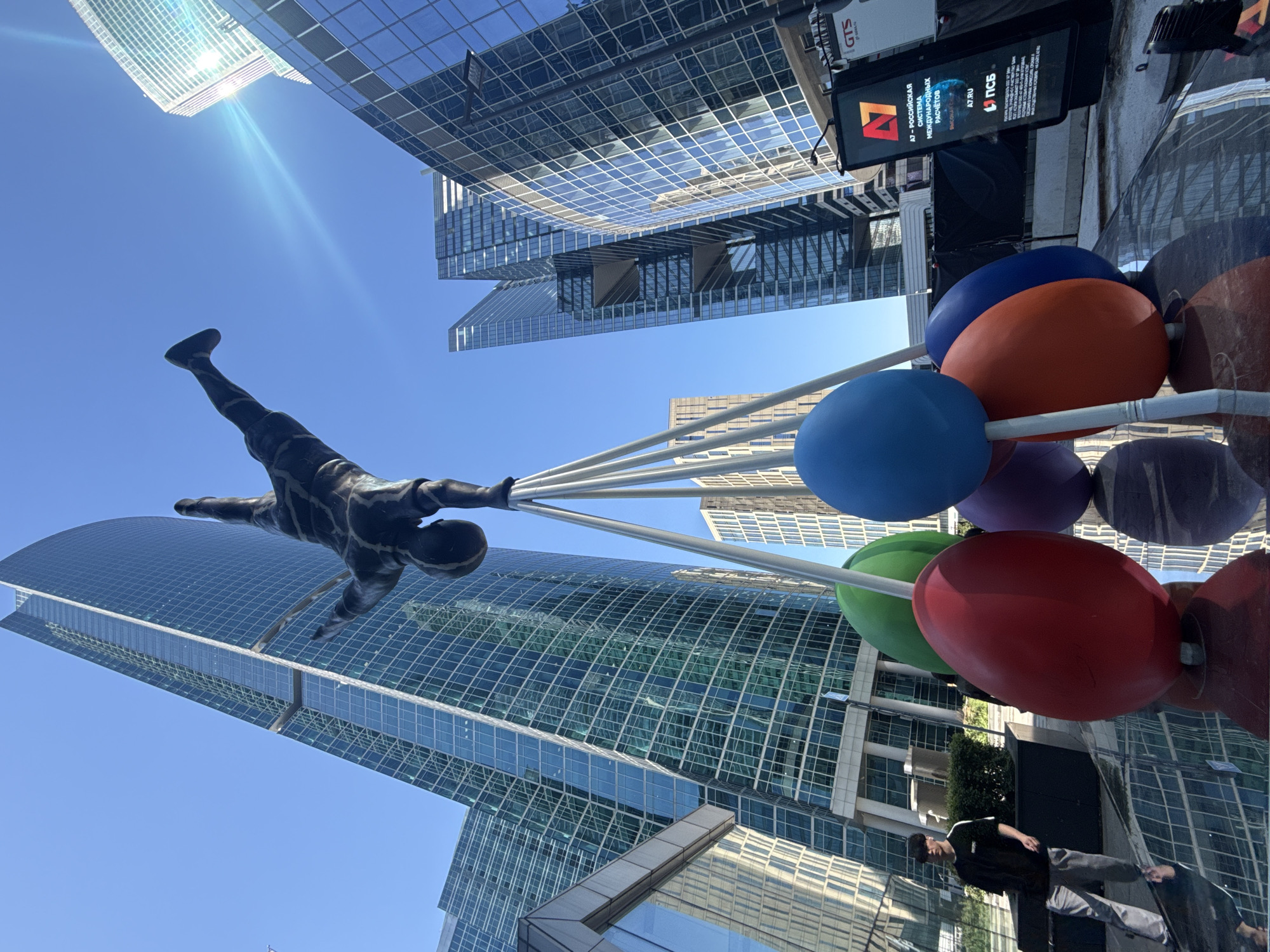 The image displays a contemporary urban scene in the city of Khimki, Russia, featuring modern architecture and a prominent sculpture under a clear blue sky. A dark, possibly bronze, statue of a person performing a handstand is centrally positioned, supported by multiple slender, light-colored poles. These poles extend downwards to a base on a reflective surface, where several large, smooth, egg-shaped or spherical objects are arranged. These objects are colored red, green, blue, purple, and orange.
The ground around the statue and colorful shapes is a dark, highly polished surface, providing clear reflections of the art installation, surrounding buildings, and a nearby pedestrian. In the background, numerous glass-clad skyscrapers define the skyline. A tall, curved building with a blue-green glass facade is prominent on the left. To the right, another skyscraper features distinct horizontal open sections within its structure. The sun is visible in the upper right, casting a lens flare across the sky.
On the lower-left side, a person is walking, dressed in a dark t-shirt with white stripes on the sleeves and light-colored pants. Their reflection is distinctly visible on the reflective ground. To the far right, a large digital display board is visible, showing text in Cyrillic that includes "A7 - РОССИЙСКАЯ СИСТЕМА МЕЖДУНАРОДНЫХ РАСЧЕТОВ" (A7 - RUSSIAN SYSTEM OF INTERNATIONAL SETTLEMENTS) and the URL "A7.RU".