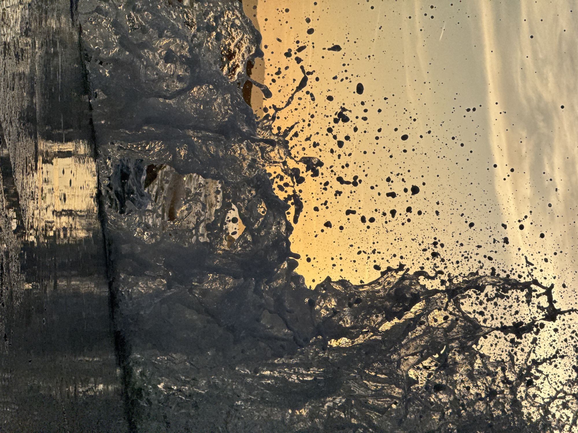 This image captures a dramatic wave crashing against the shore at sunset in Sveti Stefan, Montenegro. The foreground is dominated by the turbulent water, with splashes and droplets frozen in motion. The wave is translucent, allowing the warm, golden light of the setting sun to filter through its crest and sides. The surface of the water reflects the vibrant sky, creating a shimmering, abstract pattern. Small, dark droplets are scattered across the frame, indicating the force of the wave's impact. In the background, the sky is a gradient of orange and yellow, with faint clouds and a subtle contrail from an airplane. The setting appears to be a rocky or paved shoreline, given the hard, dark surface in the immediate foreground and its reflection. There are no people visible in the frame, the focus is entirely on the natural phenomenon of the wave and the atmospheric light.