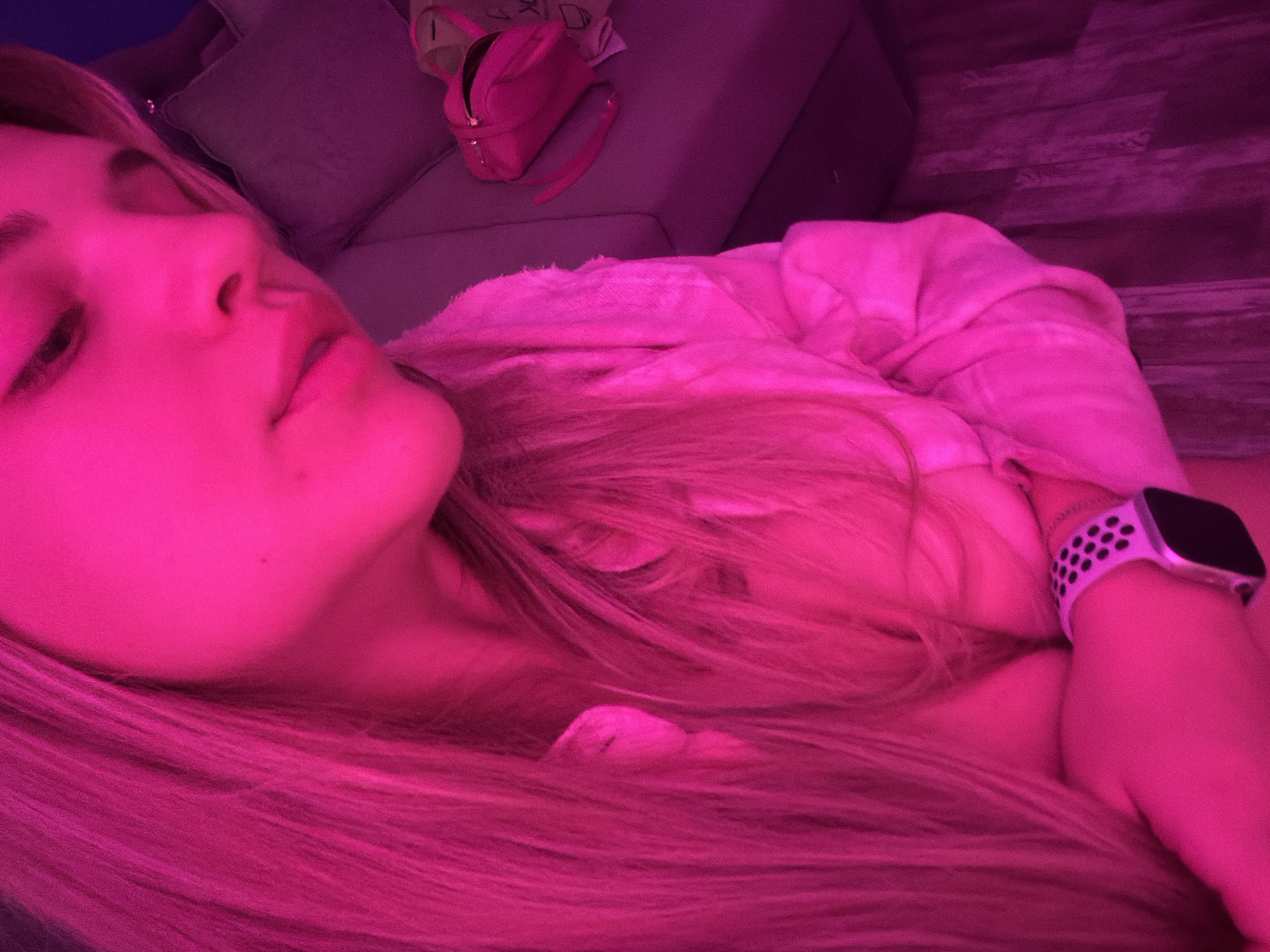 A close-up, low-angle shot shows a person lying down, likely on a couch, with a pink lighting effect dominating the scene. The person has blonde hair and is wearing a digital wristwatch with a white sport band. A pink handbag sits on the couch behind them. The location is Большая Шильна, Russia.