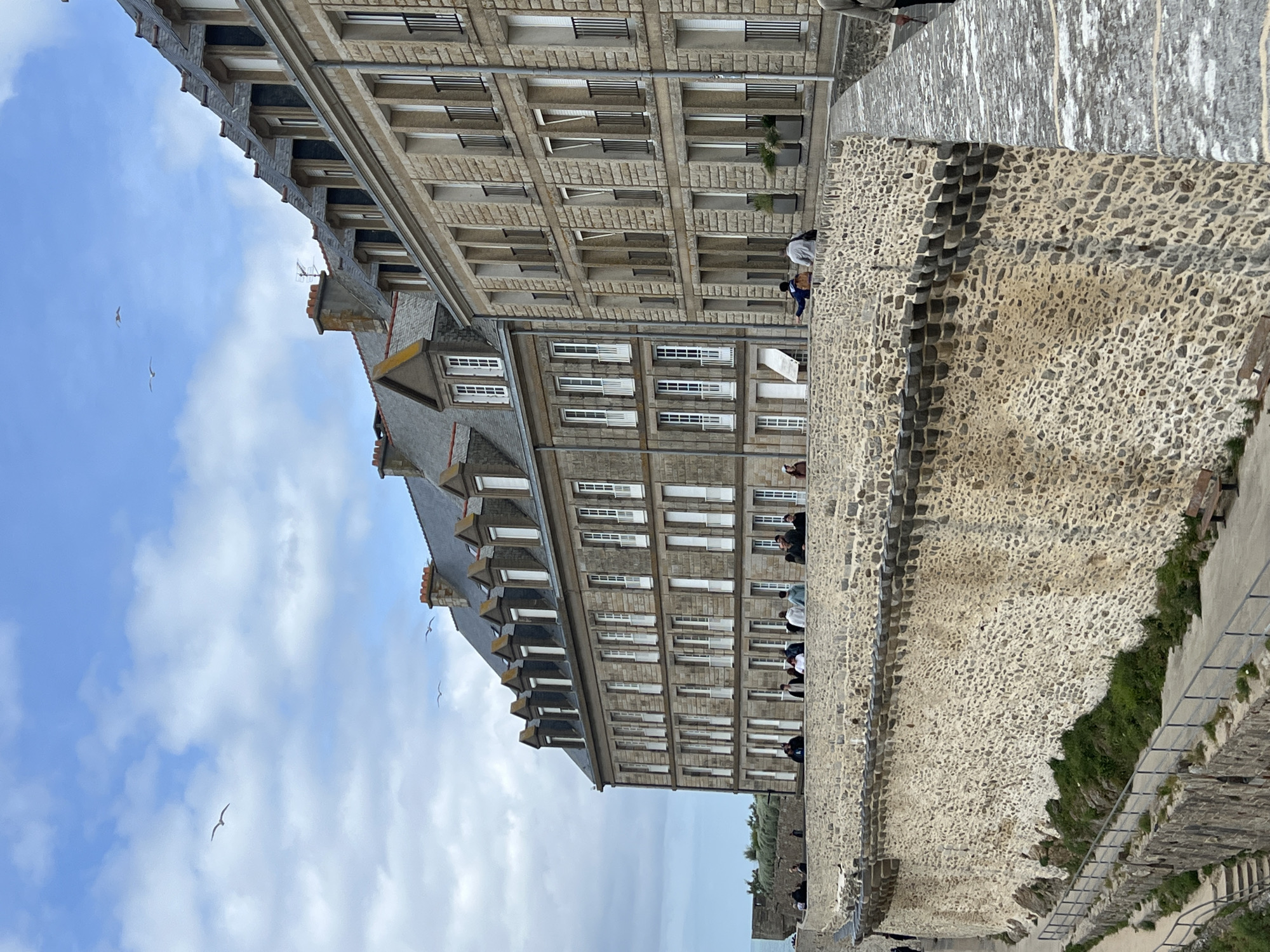 This image captures a panoramic view of the historic city of Saint-Malo, France, specifically depicting a section of its iconic ramparts and the adjacent buildings within the Intra-Muros area.
The scene is dominated by a massive, curved stone wall, which forms the foreground and mid-ground. This wall, part of the city's fortifications, is constructed from irregular, light-colored stones, with a distinct pattern of darker, stacked rectangular stones forming horizontal decorative or structural elements along its upper section. Below the main curve of the wall, a paved pathway with a dark metal railing descends gradually to the left. Patches of green vegetation are visible at the base of the wall along this lower path, where a wooden bench is also positioned.
Above this primary wall, a wider, flat stone promenade extends, where numerous people are present. These individuals, casually dressed, are observed walking, standing, and conversing in small groups. Their positions suggest they are traversing the ramparts.
Behind the promenade, two multi-story stone buildings with grey slate roofs are prominently featured. These structures exhibit a consistent architectural style, characterized by facades of light brown-grey cut stone blocks, numerous tall, narrow windows with white frames, and several dormer windows integrated into the steep roofs. The building on the right, closer to the camera, features black metal Juliet balconies on some windows and rectangular planters with greenery on its lower level. Chimneys are visible on both rooftops, and a television antenna is present on one.
The sky is overcast with a mix of light blue and scattered white and grey clouds. Several seagulls are in flight across the sky, with one prominent bird near the upper left quadrant. To the far left background, the calm, grey-blue expanse of the sea is visible, meeting the horizon under the cloudy sky, with a distant landmass or breakwater present.