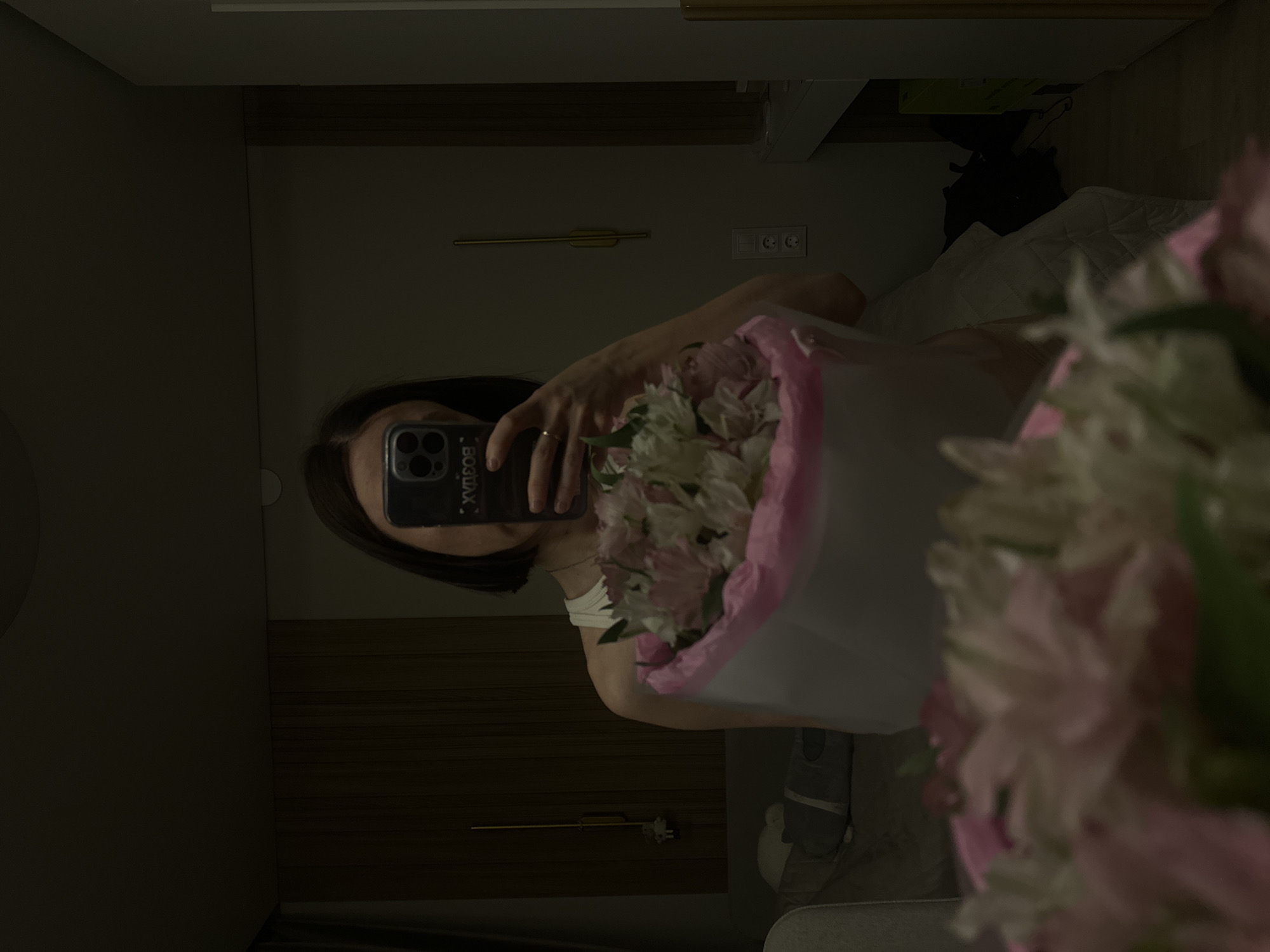A woman takes a mirror selfie while holding a bouquet of pink and white flowers. Her face is obscured by her phone. The backdrop reveals part of a bedroom with a bed, pillow, and electrical outlet. The location is likely indoors and the general context is not identifiable as Votkinsk, Russia.