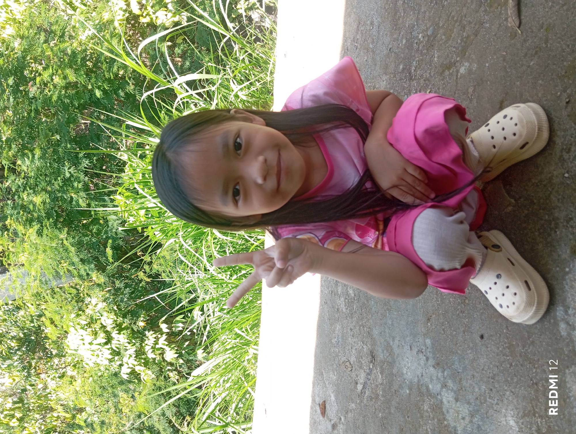 A young girl with long, dark hair is crouching on a concrete surface. She is wearing a pink t-shirt and pink skirt, white socks, and beige clogs. Her left hand is raised, with her index and middle fingers extended in a peace sign. Behind her, there is dense green foliage and tall grass, suggesting an outdoor setting. The image is labeled "REDMI 12" in the bottom left corner. This scene is likely located in a rural or suburban area of Pagar Alam, Indonesia, characterized by its abundant natural vegetation.