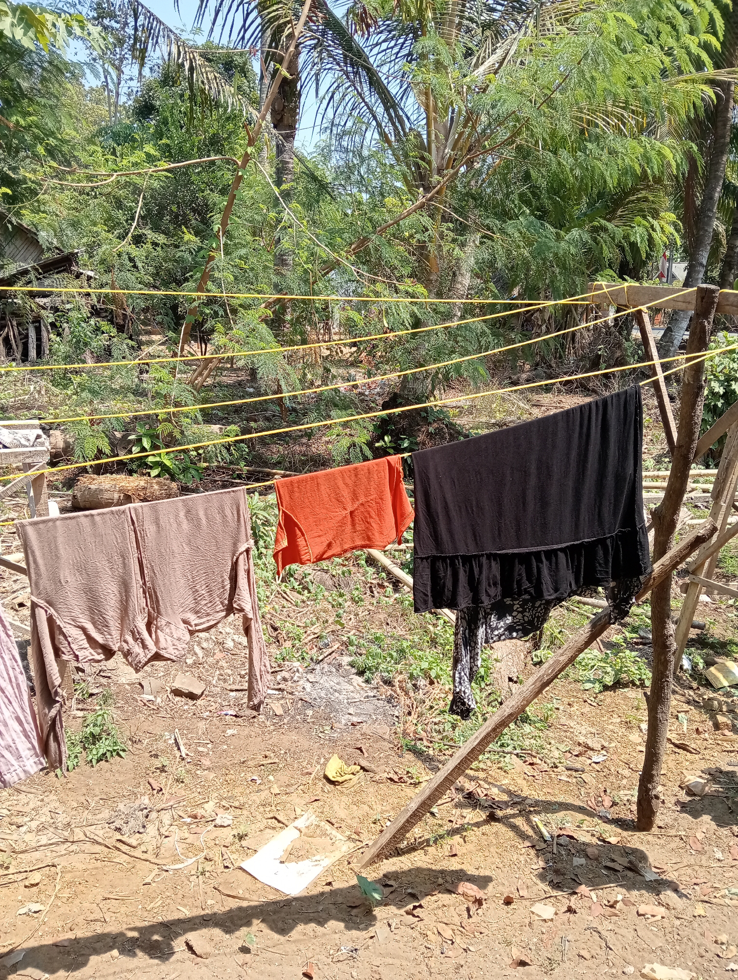 The image depicts an outdoor scene in Muara, Indonesia, featuring a clothesline strung between two wooden posts. Three garments are hanging on the line: a light brown garment on the left, an orange garment in the middle, and a black garment on the right. A partially visible pink and white striped garment is also present on the far left. The ground below is a mixture of dirt, dry leaves, and some scattered green vegetation. In the background, lush green trees and foliage are visible, suggesting a rural or semi-rural environment. Sunlight casts shadows on the ground, indicating a bright, daytime setting. The overall impression is one of domestic activity in a natural setting.
