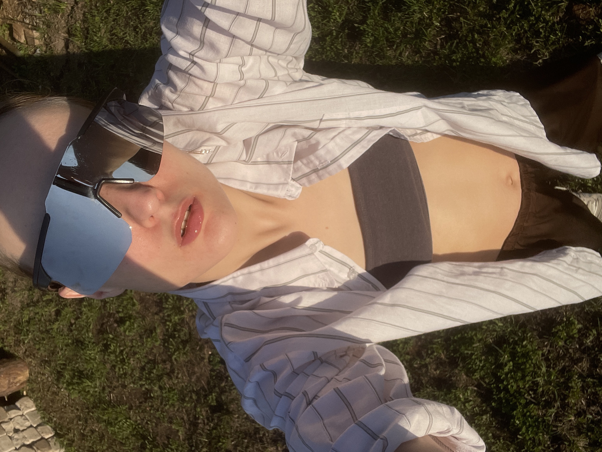 A person lies on the grass in Подшивалово, Russia, wearing silver mirrored sunglasses and a white, striped shirt that is partially unbuttoned. A dark-colored top is visible underneath the shirt. The person is also wearing brown pants. The scene is brightly lit by sunlight. A cobblestone path is visible in the bottom left corner.