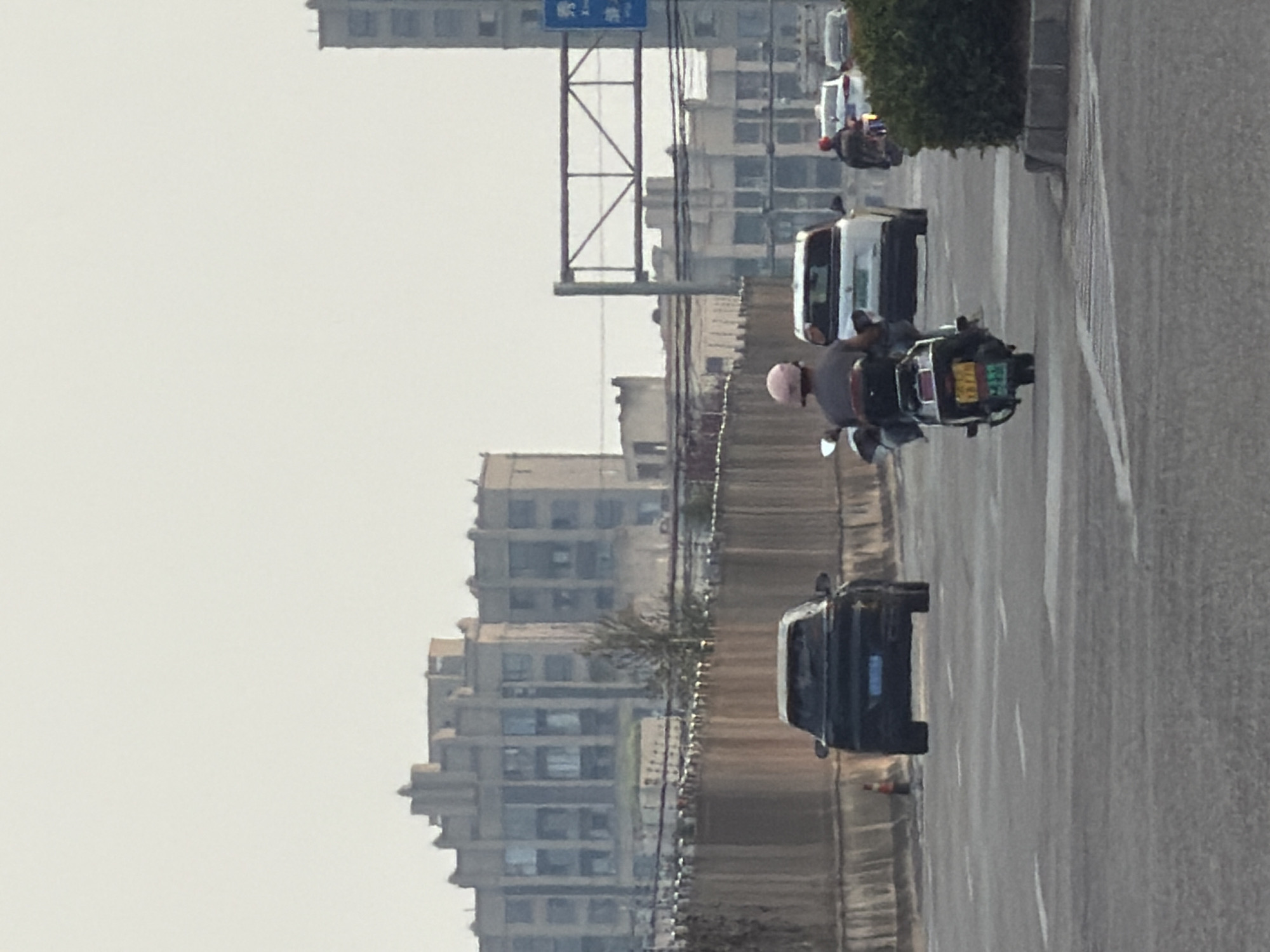 The image shows a street scene in Jiangjin, China. In the foreground, a person is riding a scooter with a passenger, both wearing helmets. To their left, a dark-colored car is driving away. Further down the road, a white SUV and a dark-colored car are visible, along with another scooter with a rider. In the background, several multi-story buildings with numerous windows are seen, along with a fence-like structure separating the road. The sky is overcast.