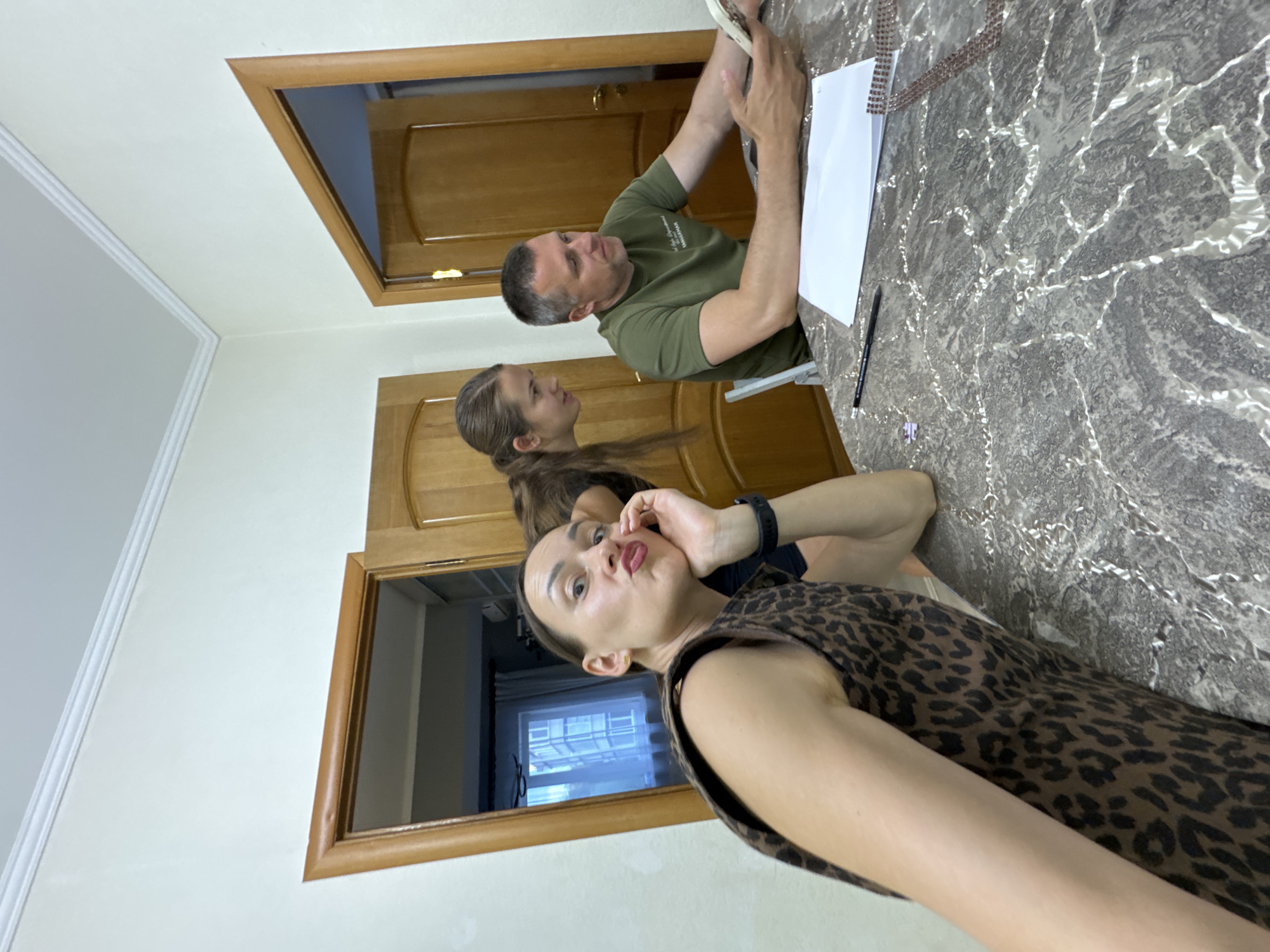 The image shows a family in what appears to be a dining room in Sochi, Russia. A woman in a leopard print top is in the foreground, making a kissing face at the camera. Behind her, a teenage girl with her hair in a ponytail leans over a table. A man in a green t-shirt is seated at the table, looking at a smartphone. The room has light-colored walls and wooden doors. A marble-patterned table is in the center of the image, with a sheet of paper and a pen on it. The overall scene suggests a casual family gathering.
